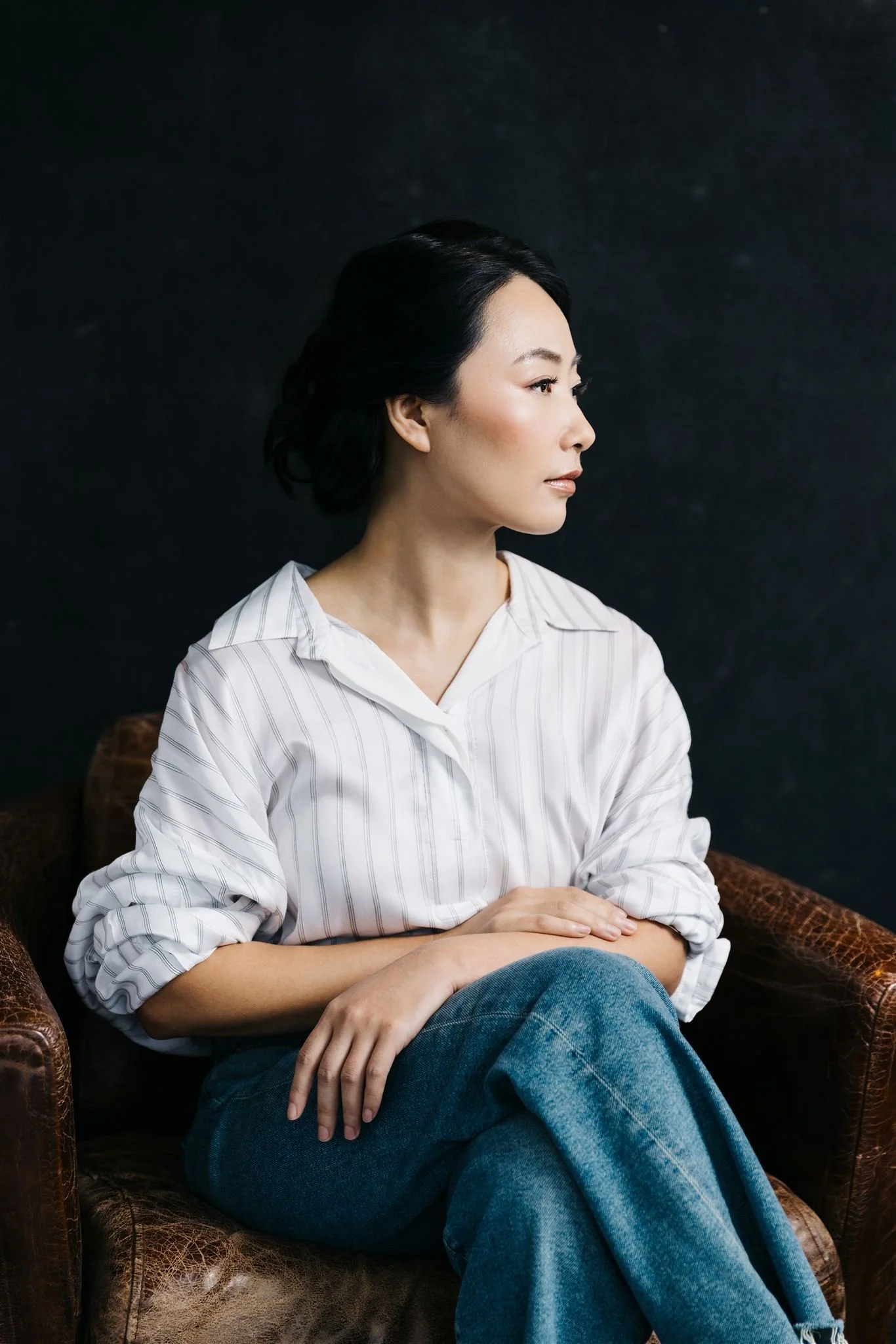 A woman with black hair, wearing a white striped shirt, sitting on a brown leather chair with dark background, looking to the right.