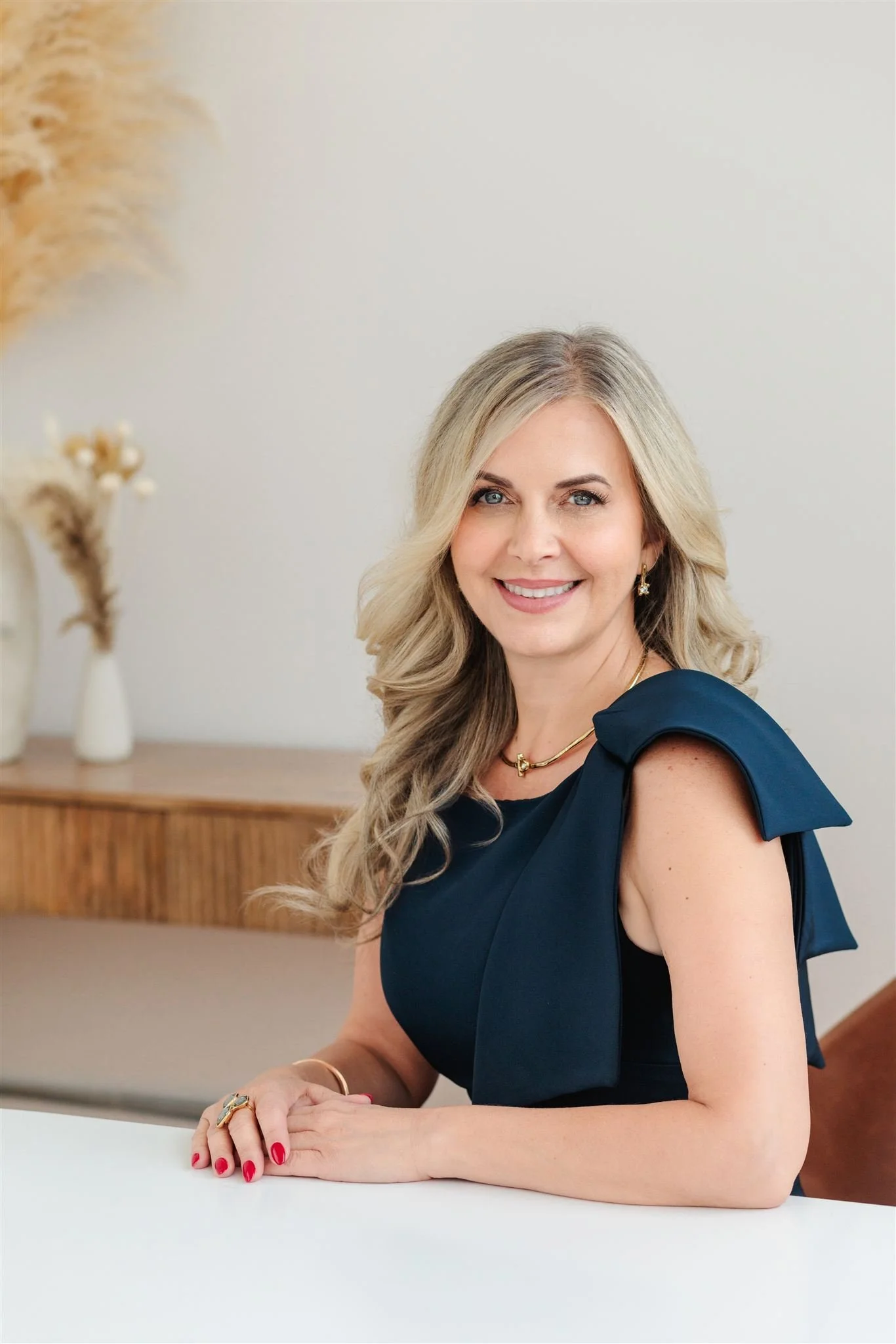 A woman with blonde, wavy hair smiling, wearing a navy blue sleeveless dress with ruffled shoulder details, sitting at a white table, with a wooden sideboard and decorative vases with dried plants in the background.