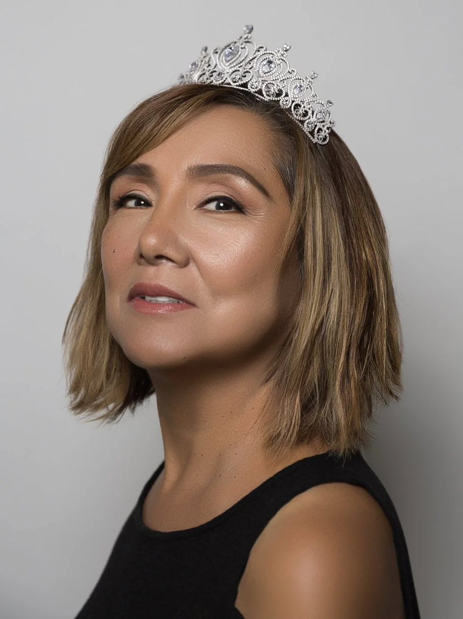 A woman with light skin and shoulder-length brown hair, wearing a silver tiara and a black sleeveless top, posing against a neutral background.
