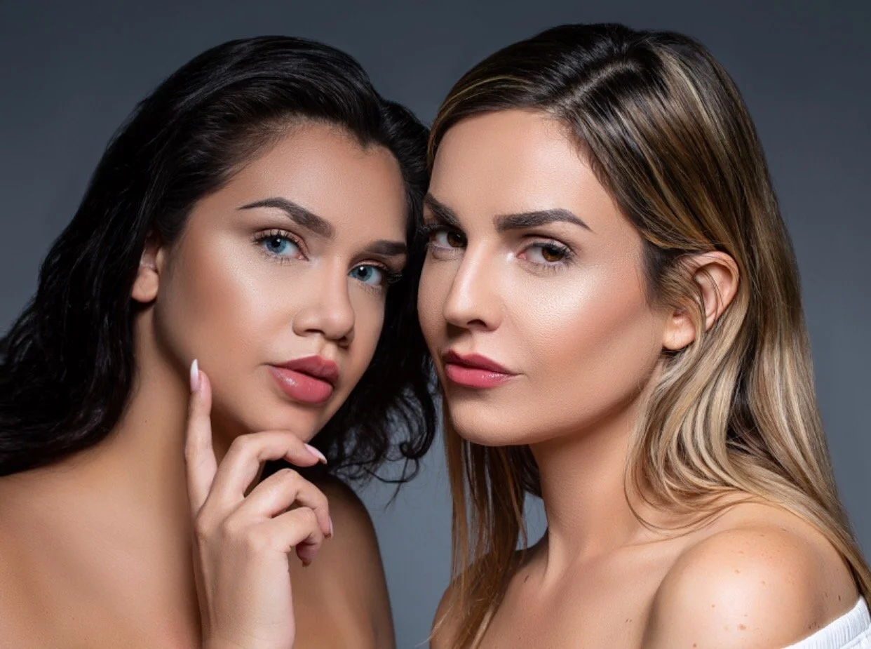 Two women with styled hair and makeup posing closely for a photo against a gray background.