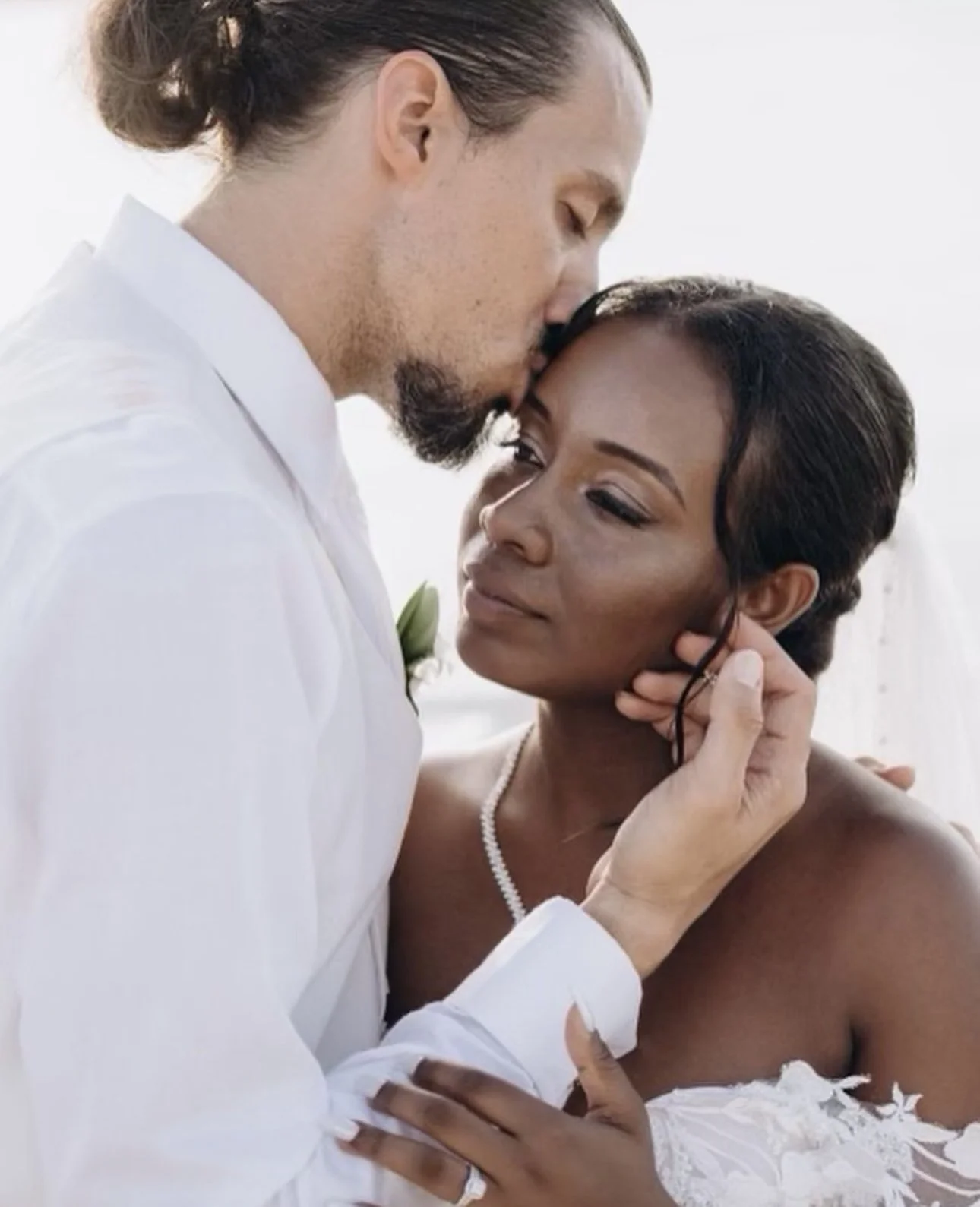A couple in wedding attire sharing an intimate moment, with the man gently kissing the woman on her head, and the woman resting her hand on his chest.