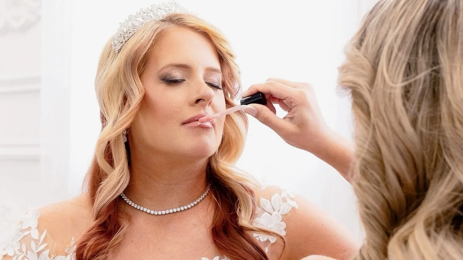 A woman with long, wavy blonde hair in a wedding dress with lace details, receiving makeup application for her lips using a lip gloss brush from a makeup artist in a bright room.
