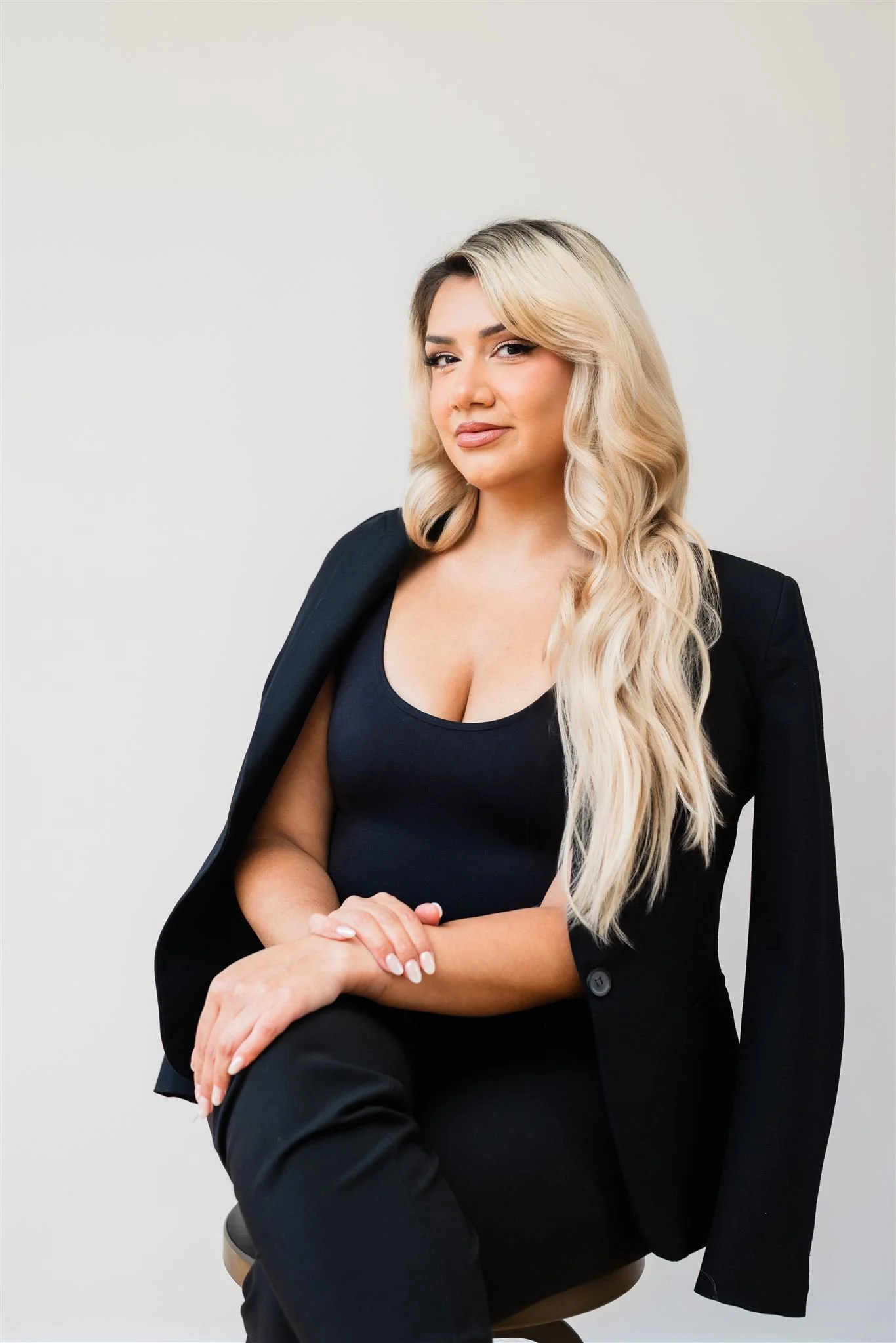 A woman with long blonde hair, wearing a black blazer over a black top, sitting against a plain light-colored background.