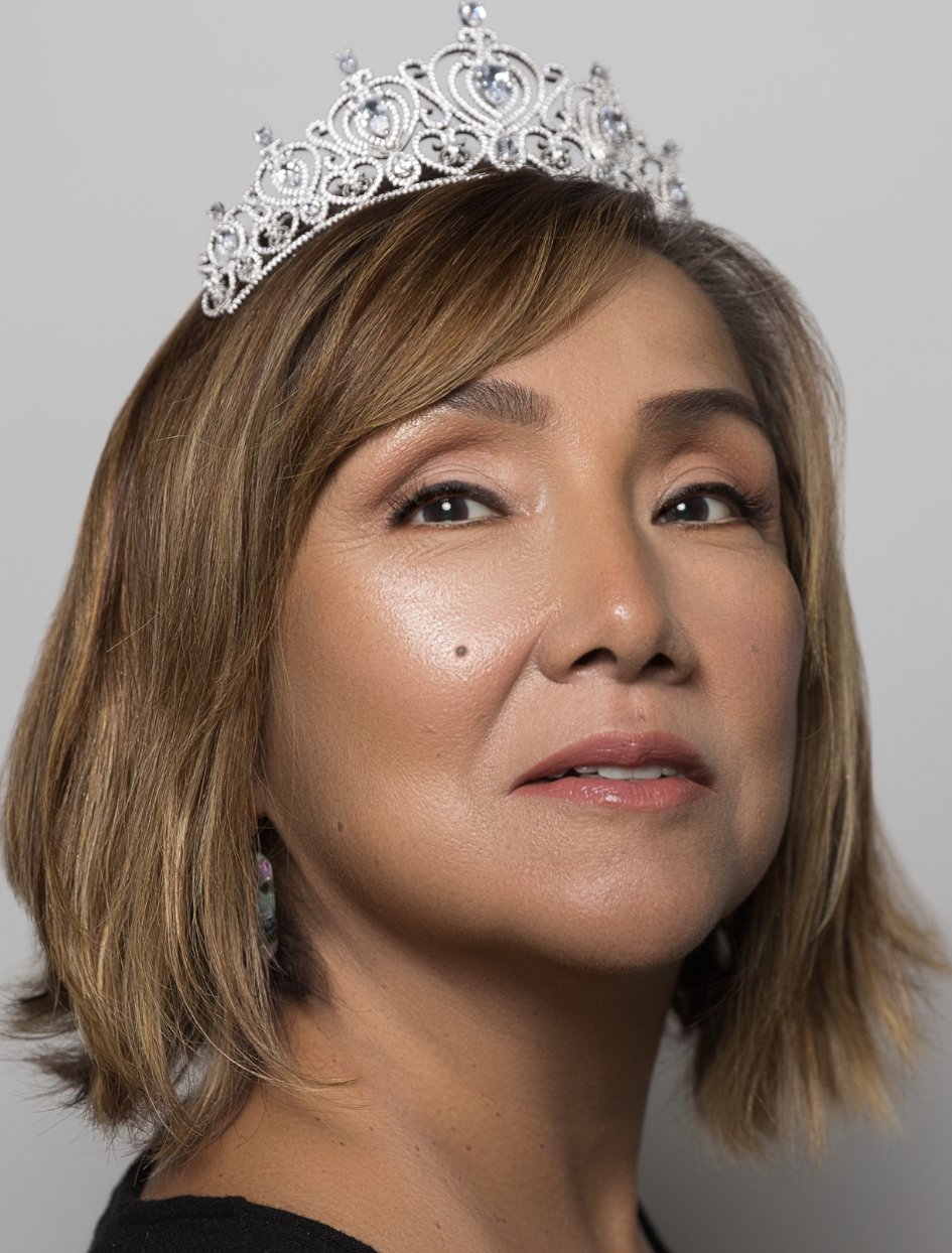 A woman with shoulder-length light brown hair wearing a silver tiara and earrings, looking slightly to the side, with a neutral facial expression.