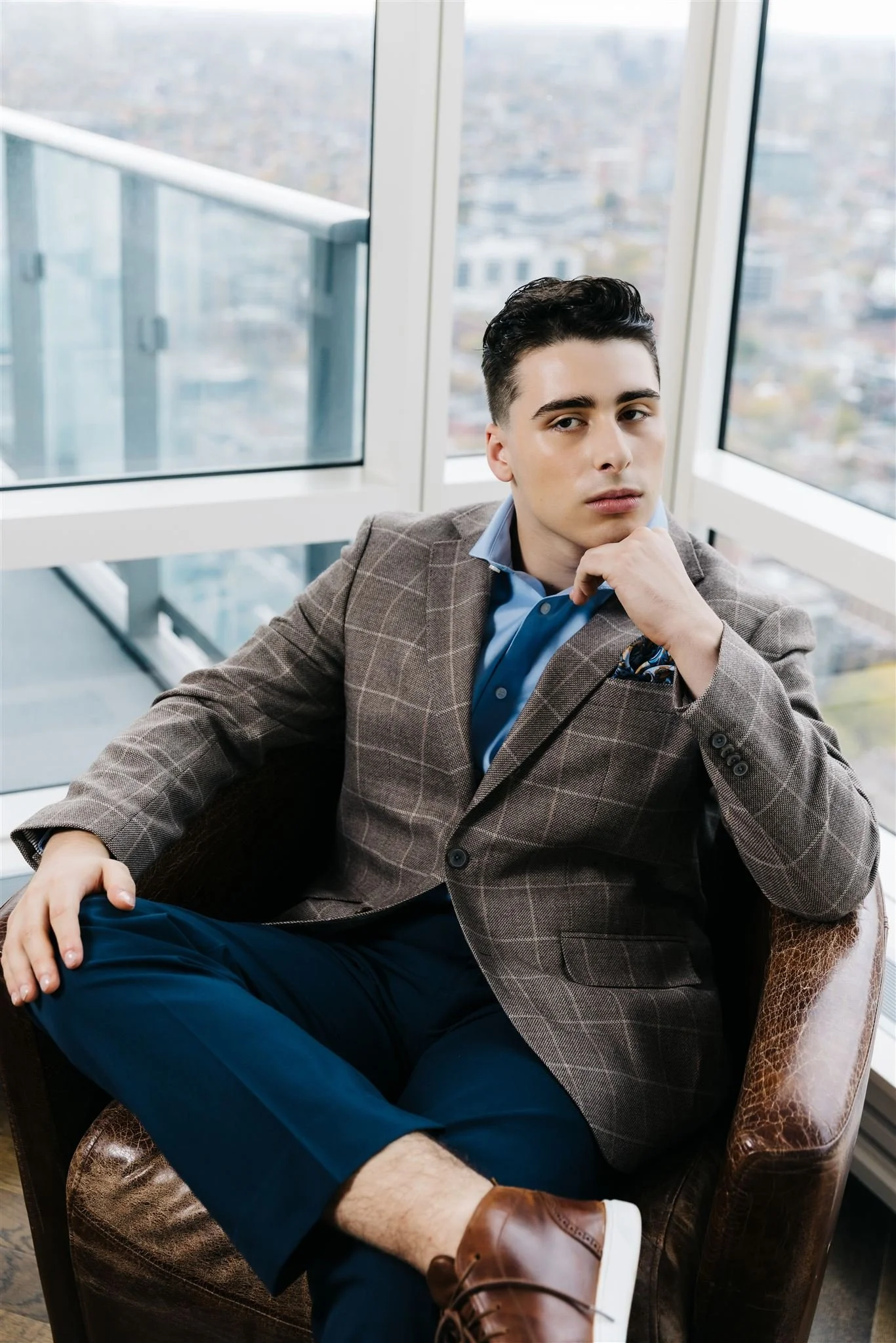 A young man with dark hair, wearing a checkered blazer and blue shirt, sitting in a leather chair by large windows overlooking a cityscape.