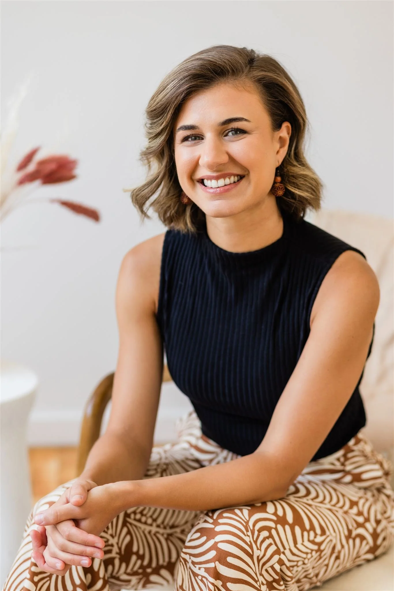 A smiling woman with wavy brown hair, wearing a sleeveless black top and patterned beige and white pants, sitting indoors near a white wall with a blurred plant in the foreground.