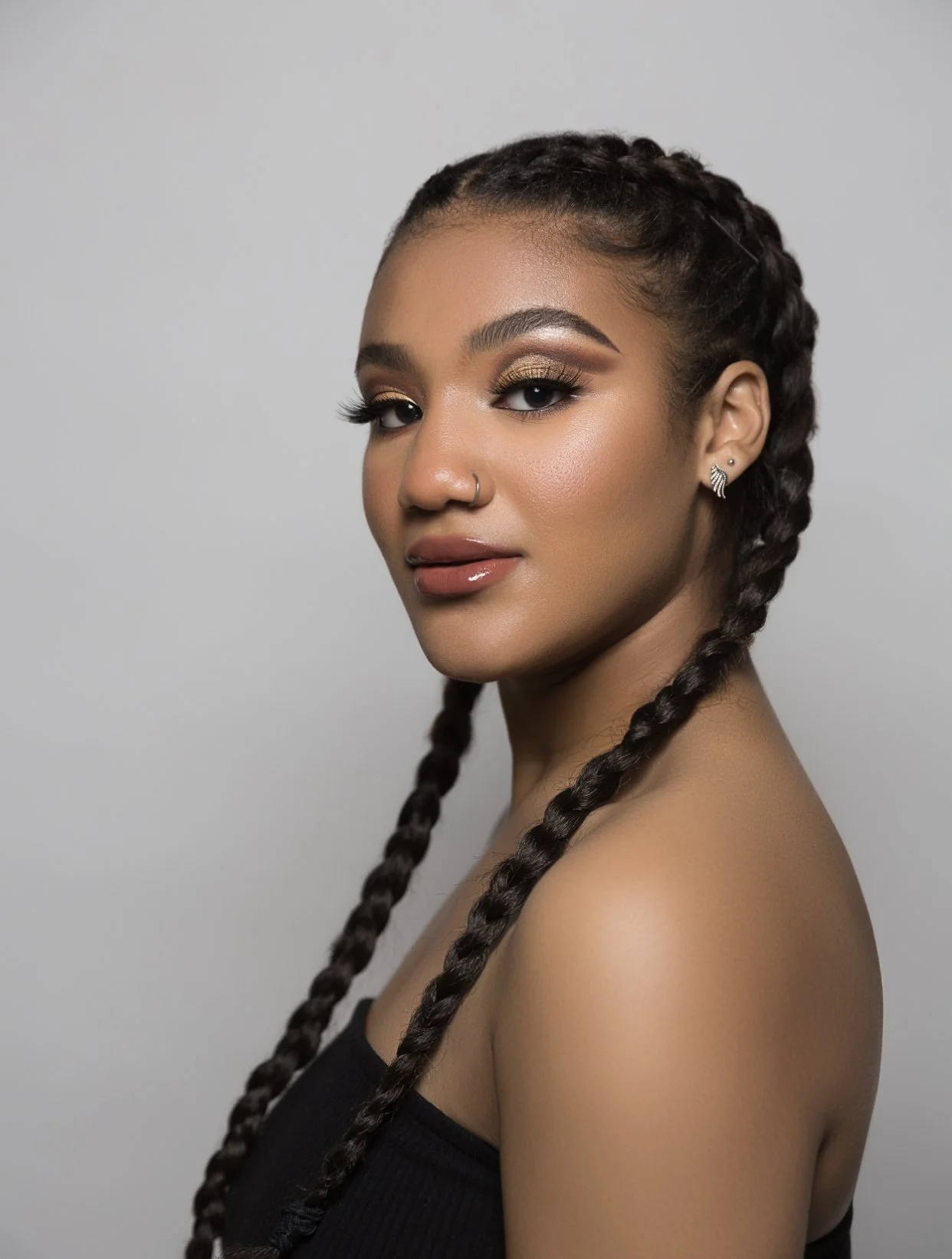 Portrait of a young woman with braided hair, gold eye makeup, and earrings, against a plain gray background.