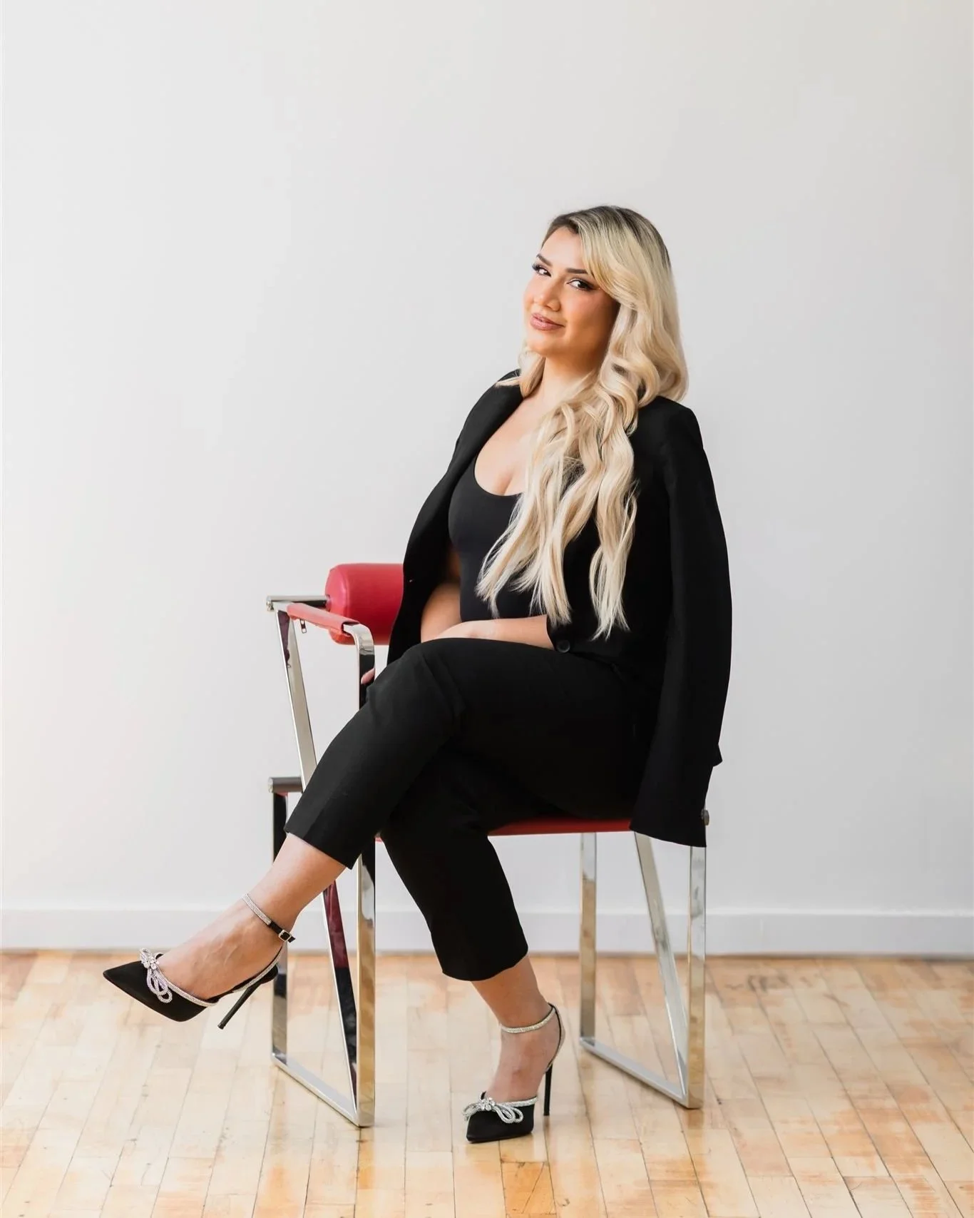 A woman with long blonde wavy hair sitting on a modern red chair with metal legs, wearing a black blazer, black top, black pants, and black high heels with white bows, against a plain white wall and hardwood floor.