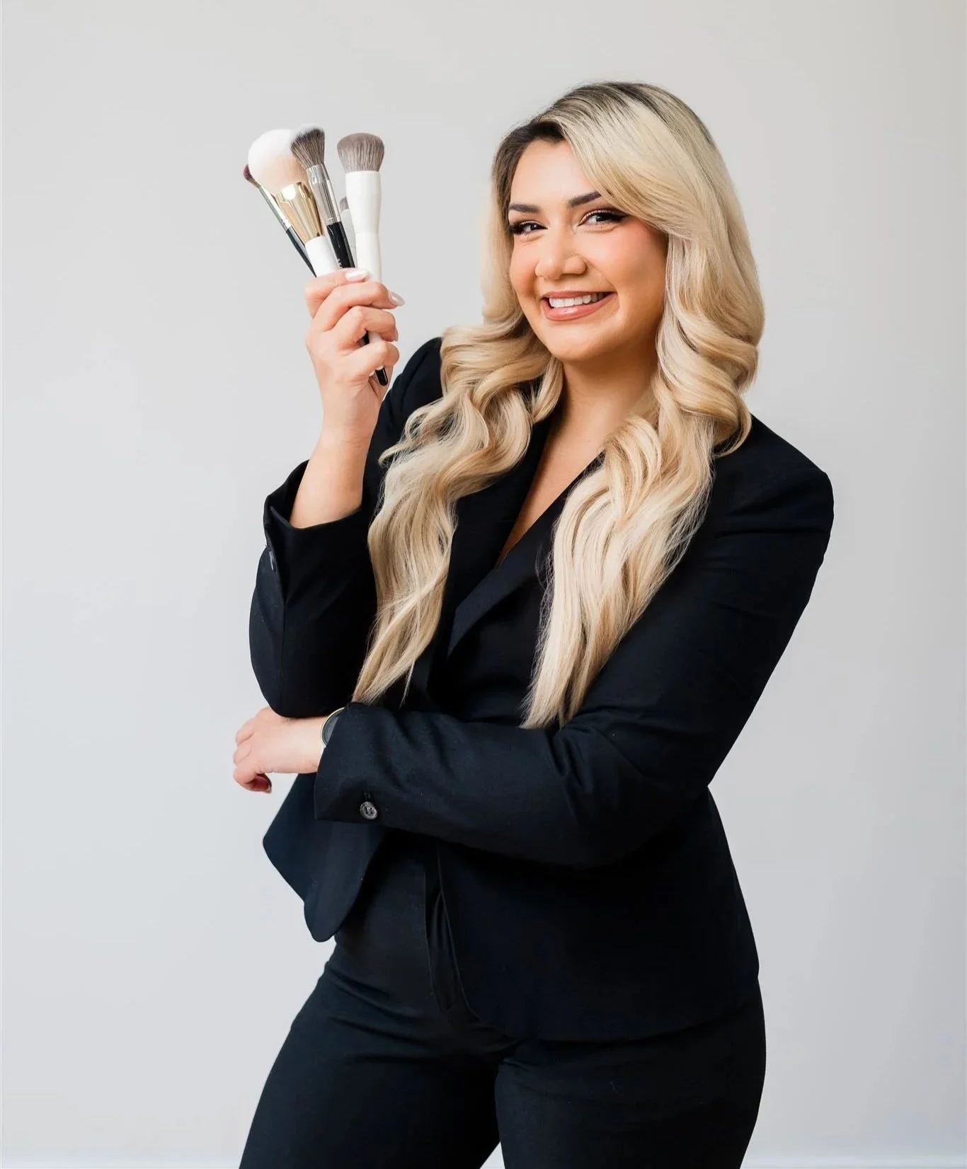 A woman with long blonde hair in loose waves, wearing a black blazer, holding five makeup brushes and smiling at the camera against a plain light gray background.