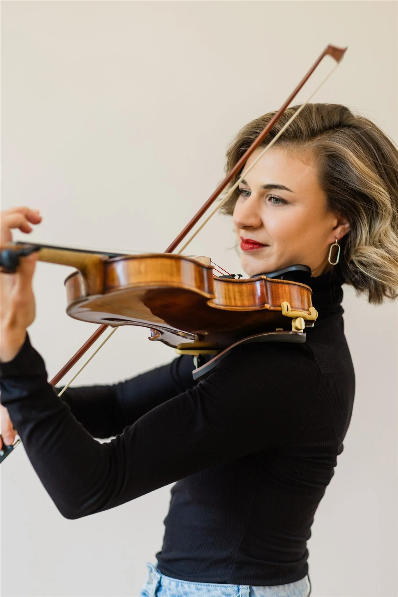 A woman with short brown hair playing a violin, wearing a black long-sleeve top and earrings, against a plain light-colored background.