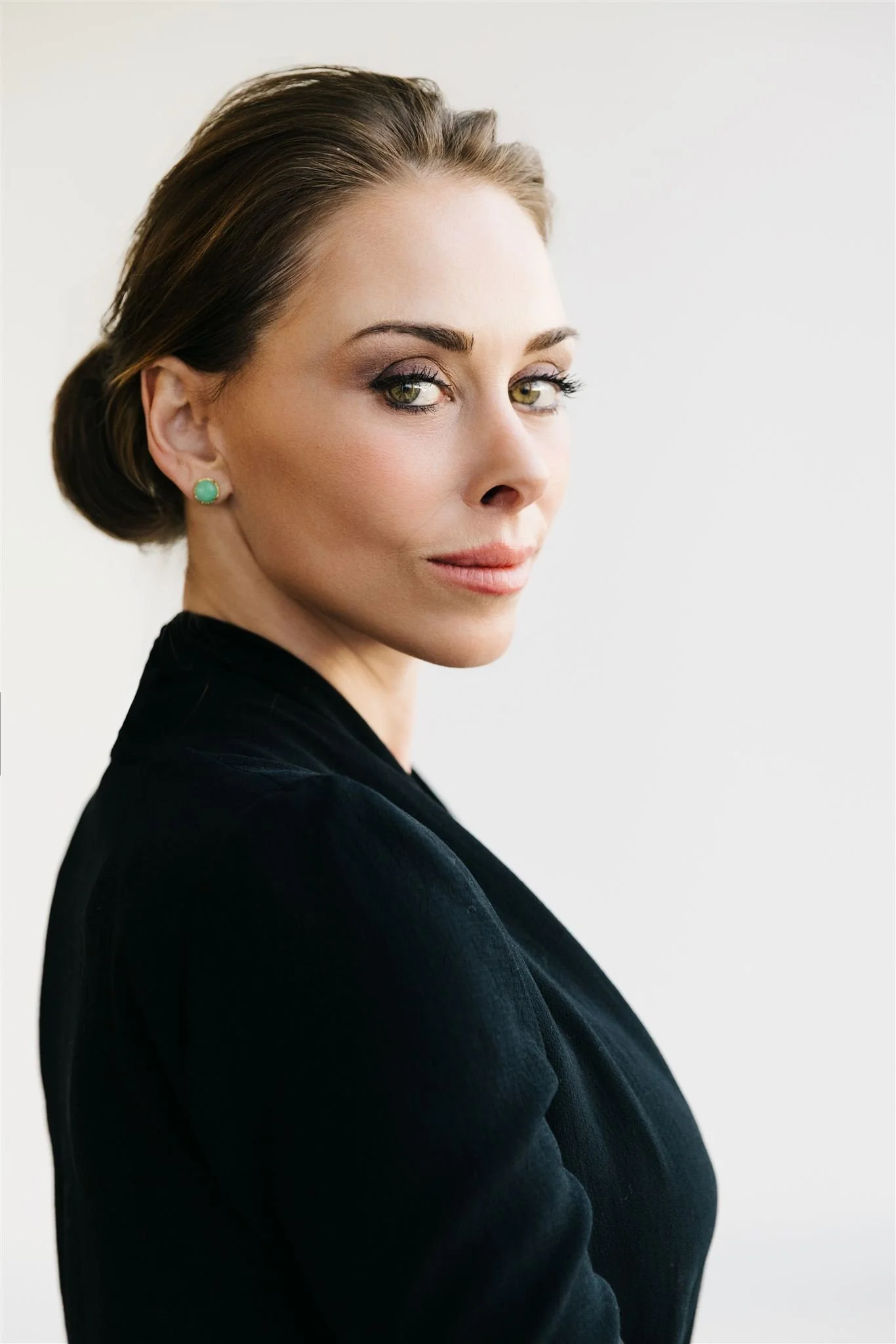 A woman with styled brown hair, wearing green earrings and minimal makeup, looking over her shoulder at the camera against a plain light background.