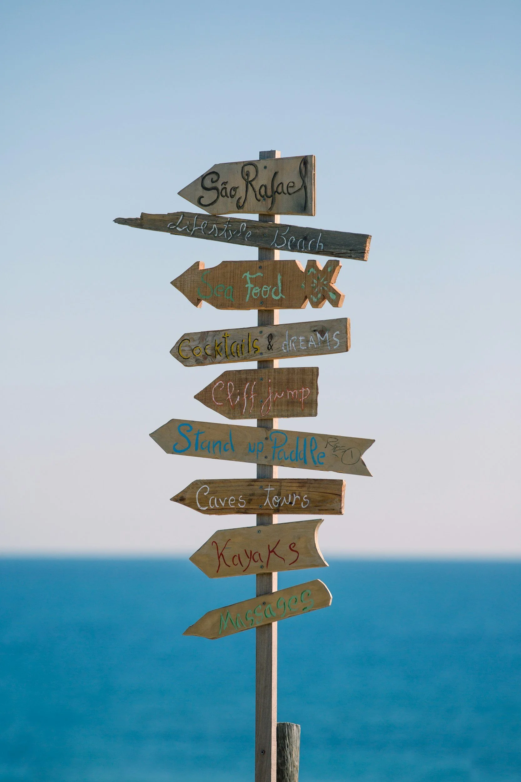 Wooden signpost with multiple directional arrows, each labeled with activities or locations like 'São Rafael,' 'Lifeguard Beach,' 'Sea Food,' 'Cocktails & dreams,' 'Cliff jump,' 'Stand up Paddle,' 'Caves Tours,' 'Kayaks,' and 'Massages,' set against a backdrop of the ocean and a clear sky.