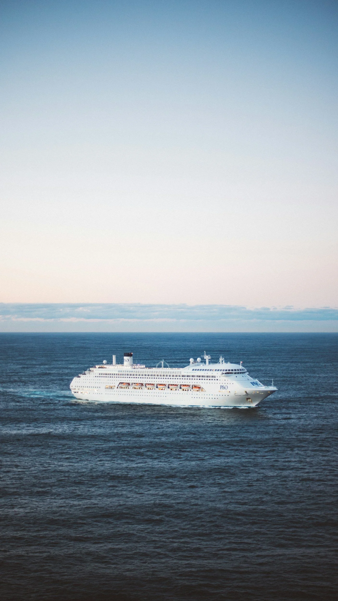 Large white cruise ship sailing on calm ocean waters with a pale blue sky in the background.