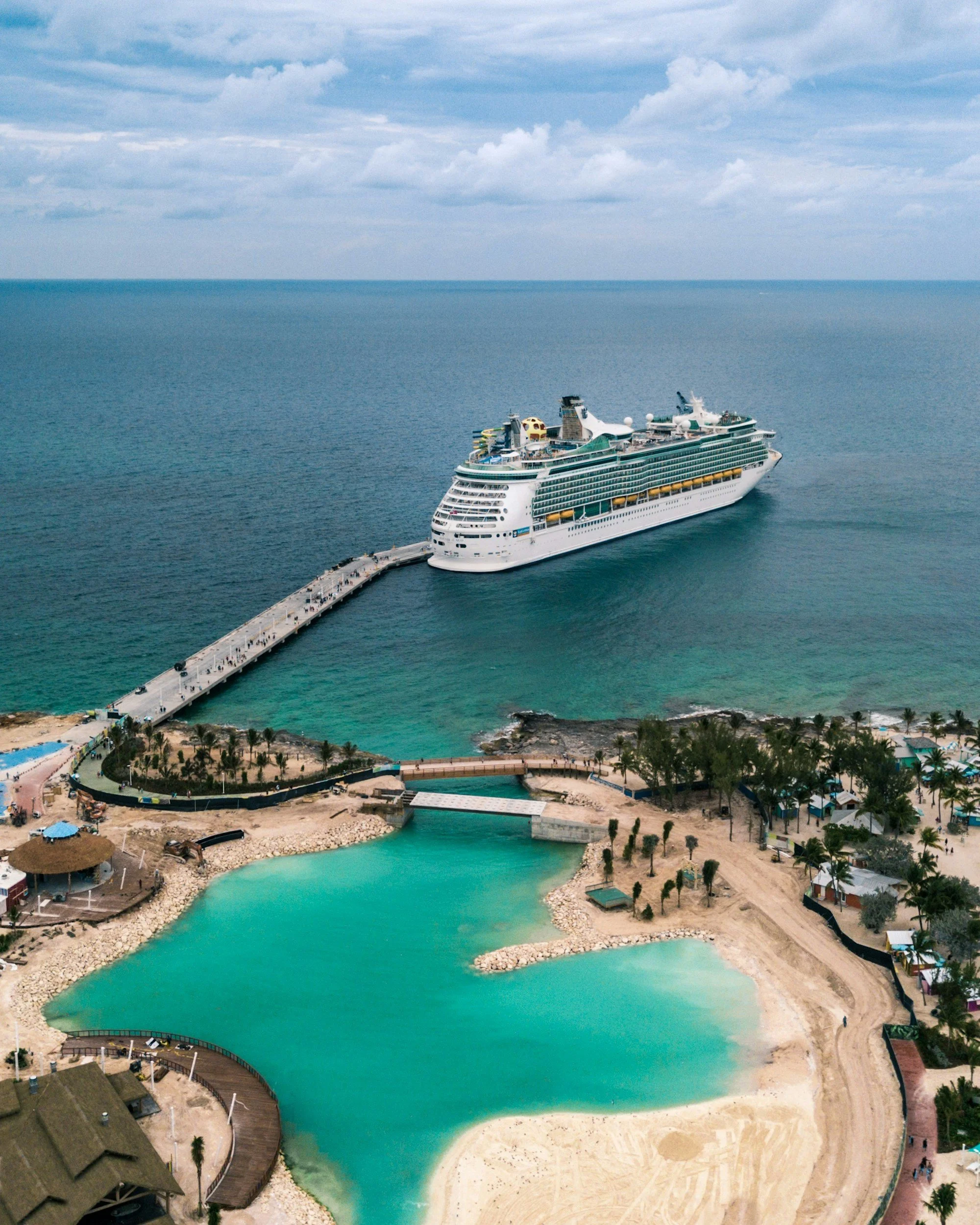 A large cruise ship docked at a pier near an artificial lagoon with palm trees, small buildings, and sandy areas.