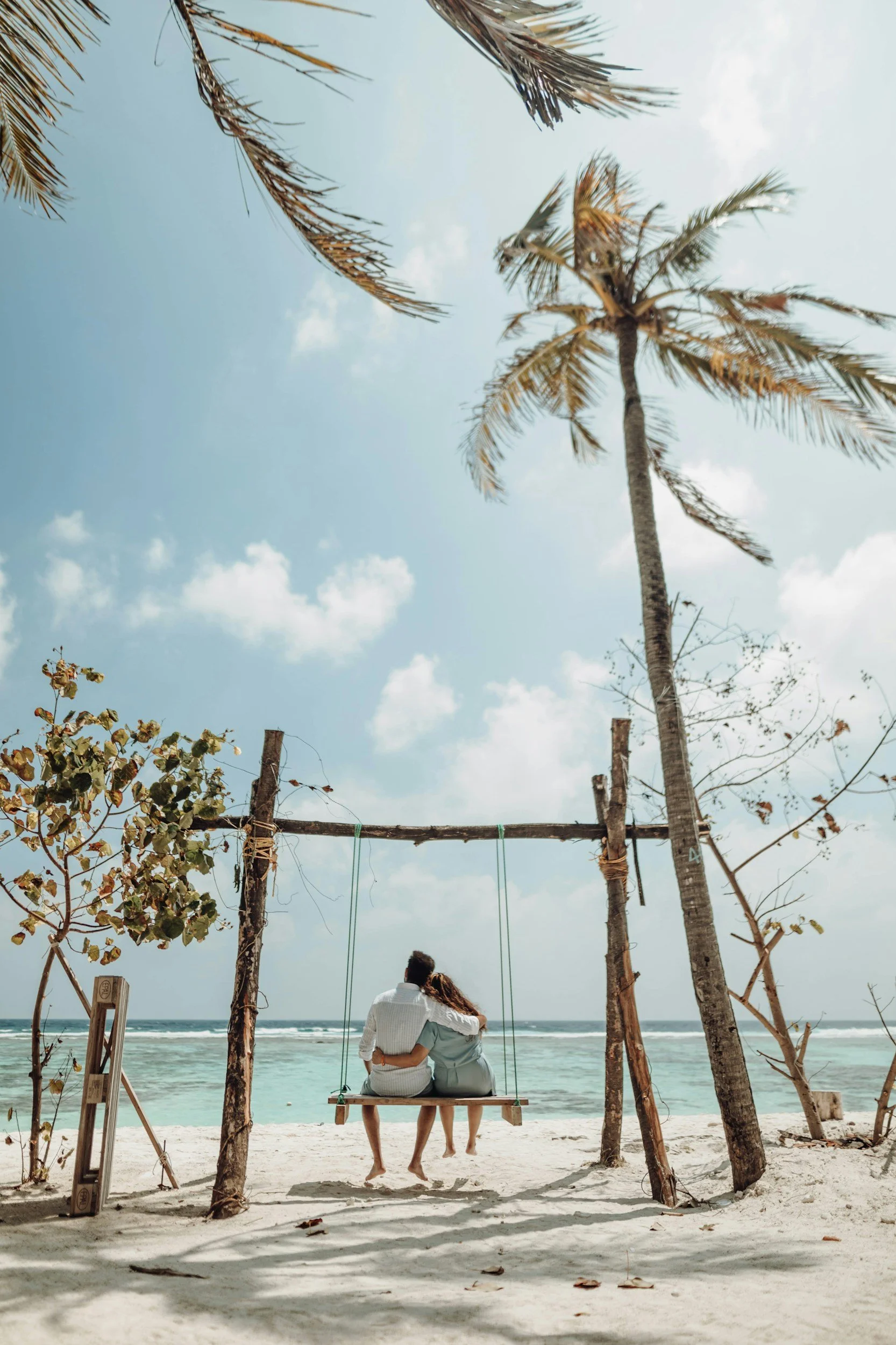 Couple sitting on a swing on a beach, overlooking the ocean with palm trees and a partly cloudy sky.