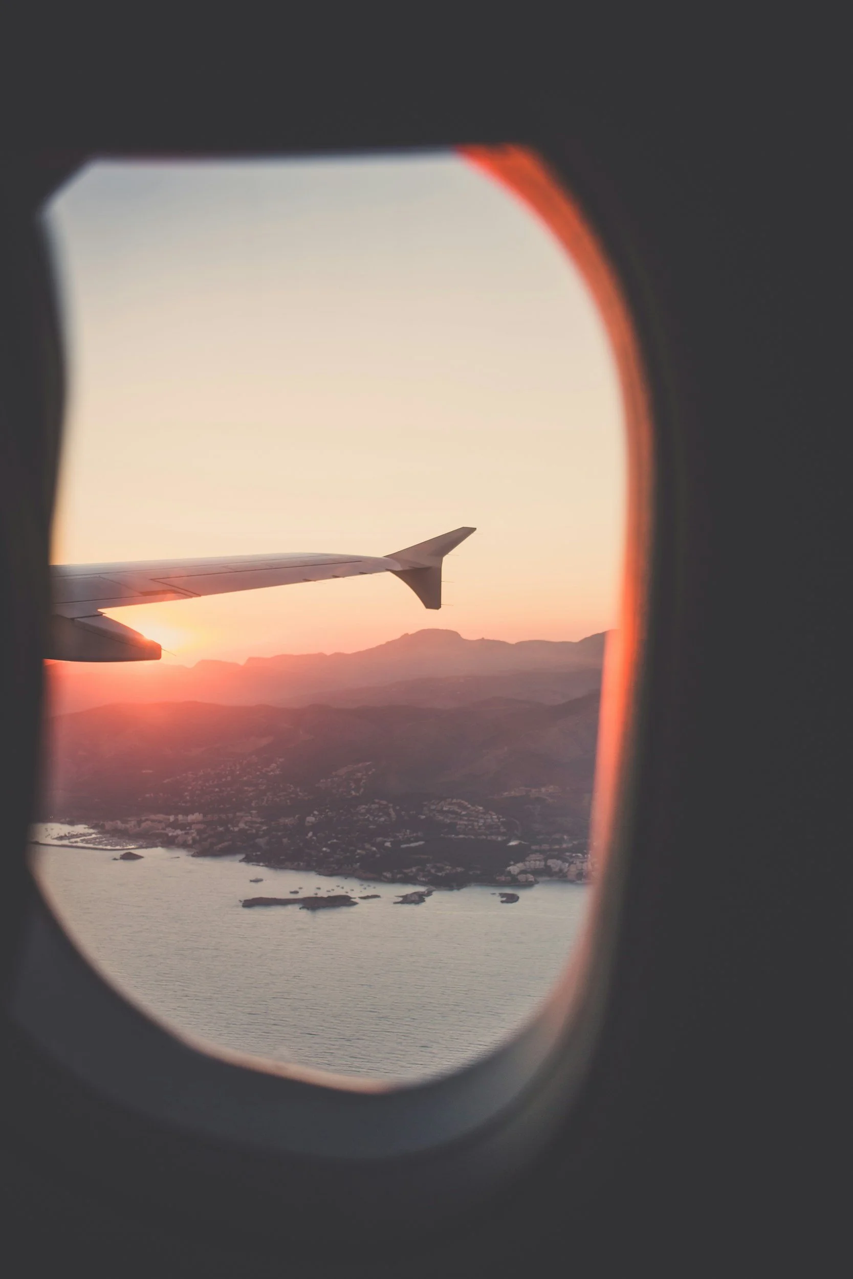View from an airplane window showing an airplane wing at sunset over a mountainous coastline with water.