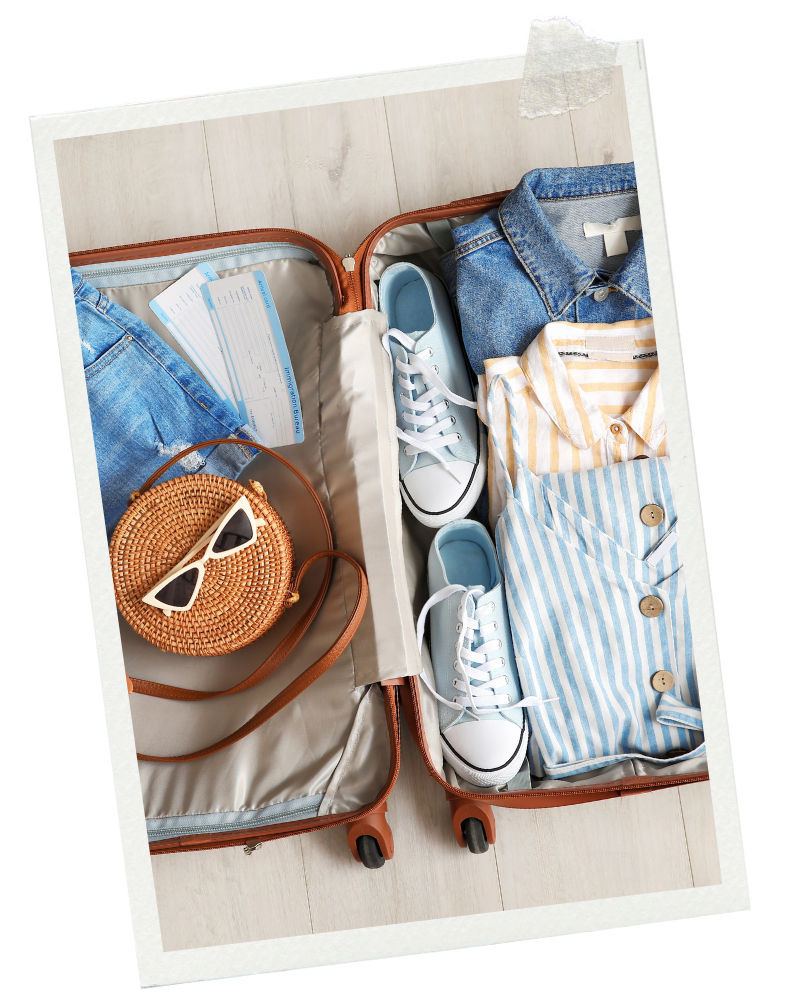 Open suitcase packed with blue and striped clothing, white sneakers, a round woven bag with sunglasses, and travel documents.