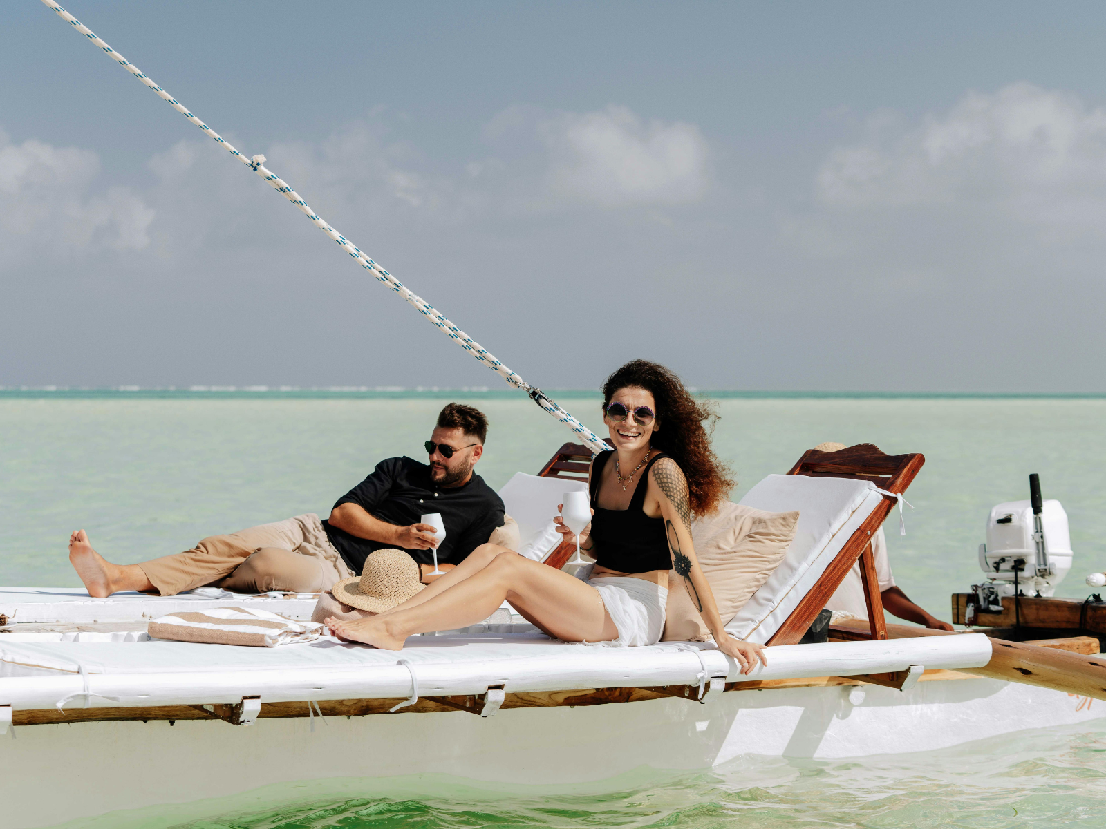 Two people relaxing on a boat in a tropical location with clear water, one woman sitting on the boat's edge and a man lying back, both smiling and holding drinks, with the ocean and sky in the background.