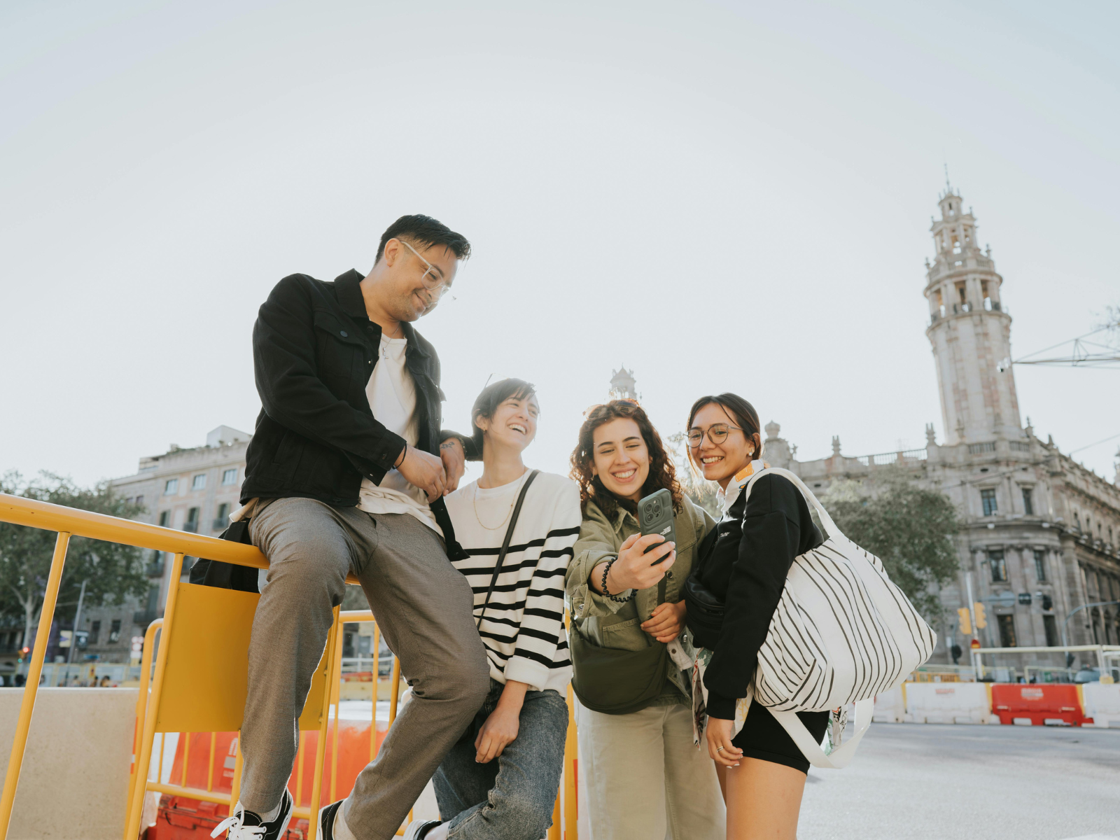 Four friends taking a selfie together outdoors, in front of a historic building and a clock tower, smiling and enjoying their time.