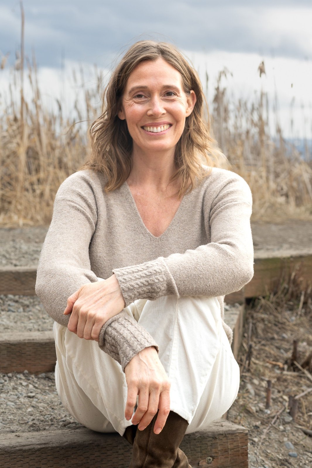 A woman with shoulder-length wavy brown hair, smiling, sitting outdoors on wooden steps with dried tall grasses behind her and cloudy sky.