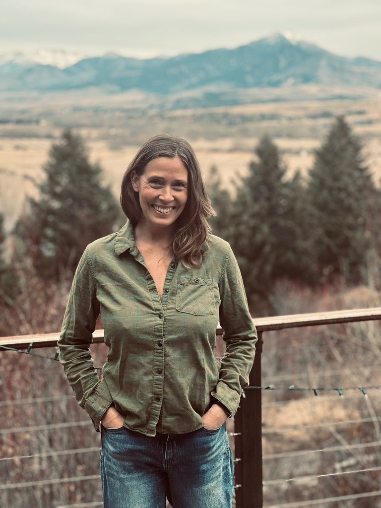 A woman with brown hair, wearing a green button-up shirt and blue jeans, standing on a wooden deck with her hands in her pockets, smiling, with trees and mountains in the background.