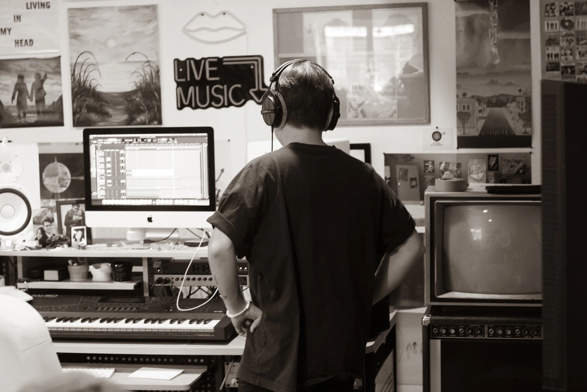 A person in headphones stands in a music studio with a computer, keyboard, and vintage TV. The room is decorated with posters and artwork, including a 'Live Music' sign and a lips drawing. The person is viewed from the back, focusing on music production.