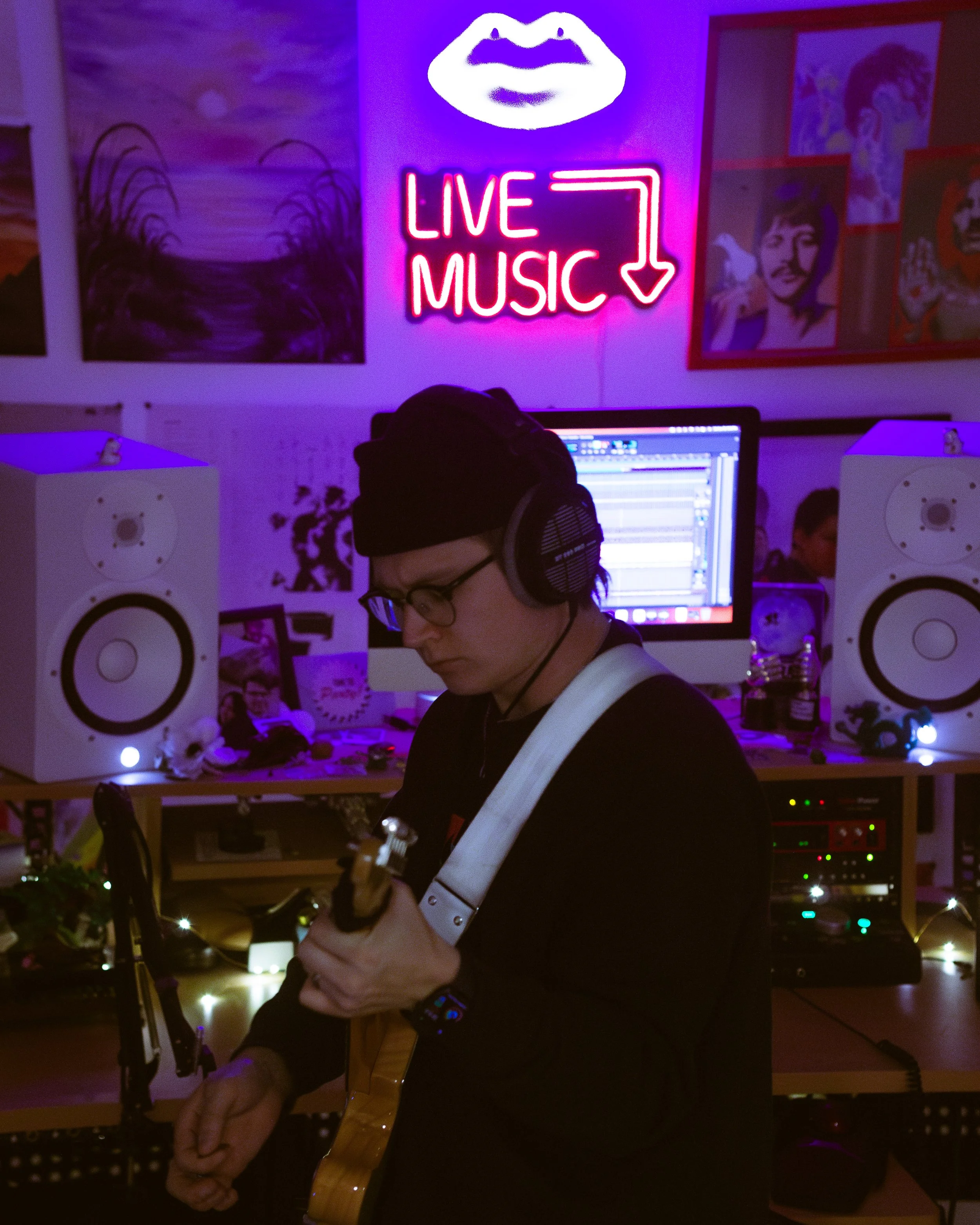 A person wearing headphones and glasses playing an electric guitar in a dimly lit room with neon signs that read 'Live Music' and a lips symbol. The room has speakers, a computer, and artwork on the walls.
