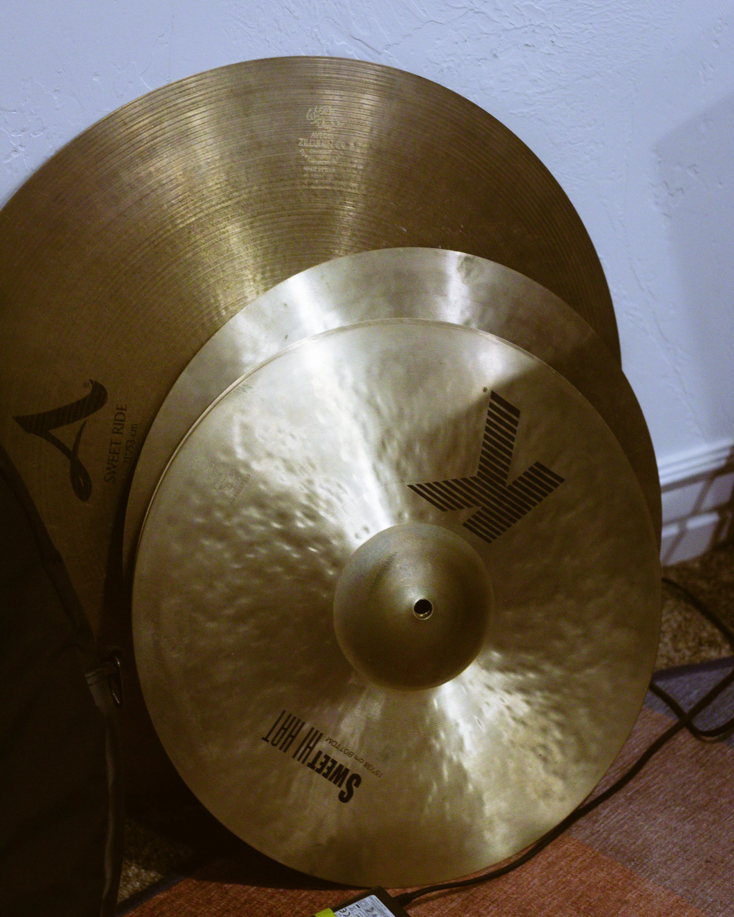 Two cymbals stacked, one larger gold-colored with a textured surface and branding, and the smaller silver-colored one on top, near a wall.