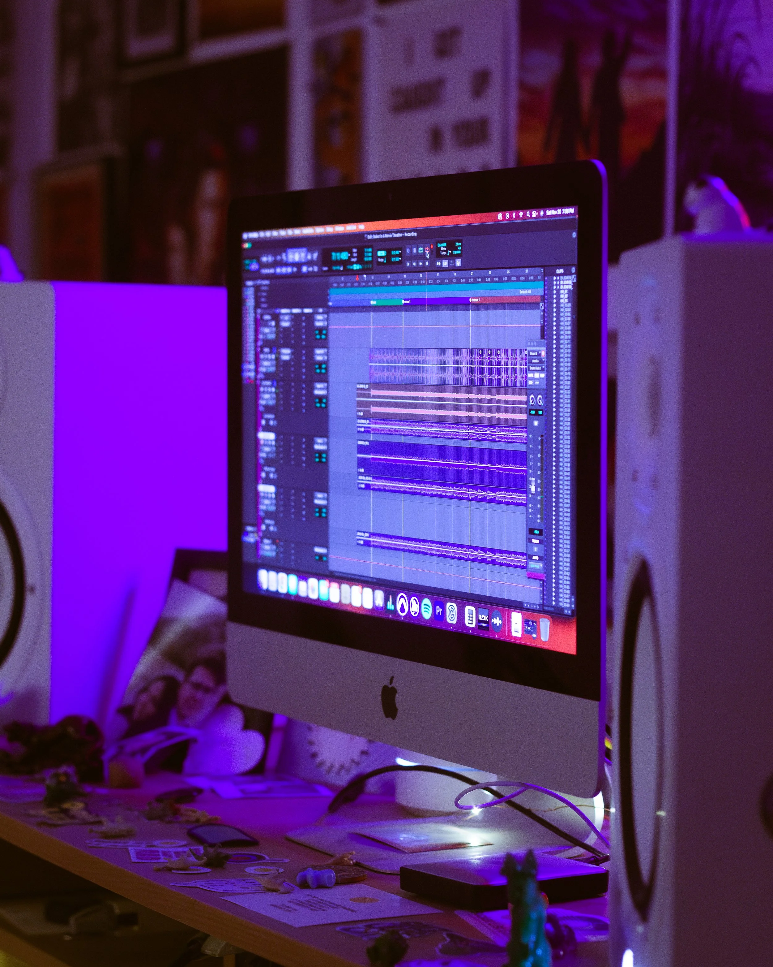 A computer monitor displaying a digital audio workstation with music tracks and waveforms, surrounded by studio speakers and decorative items in a dimly lit room.
