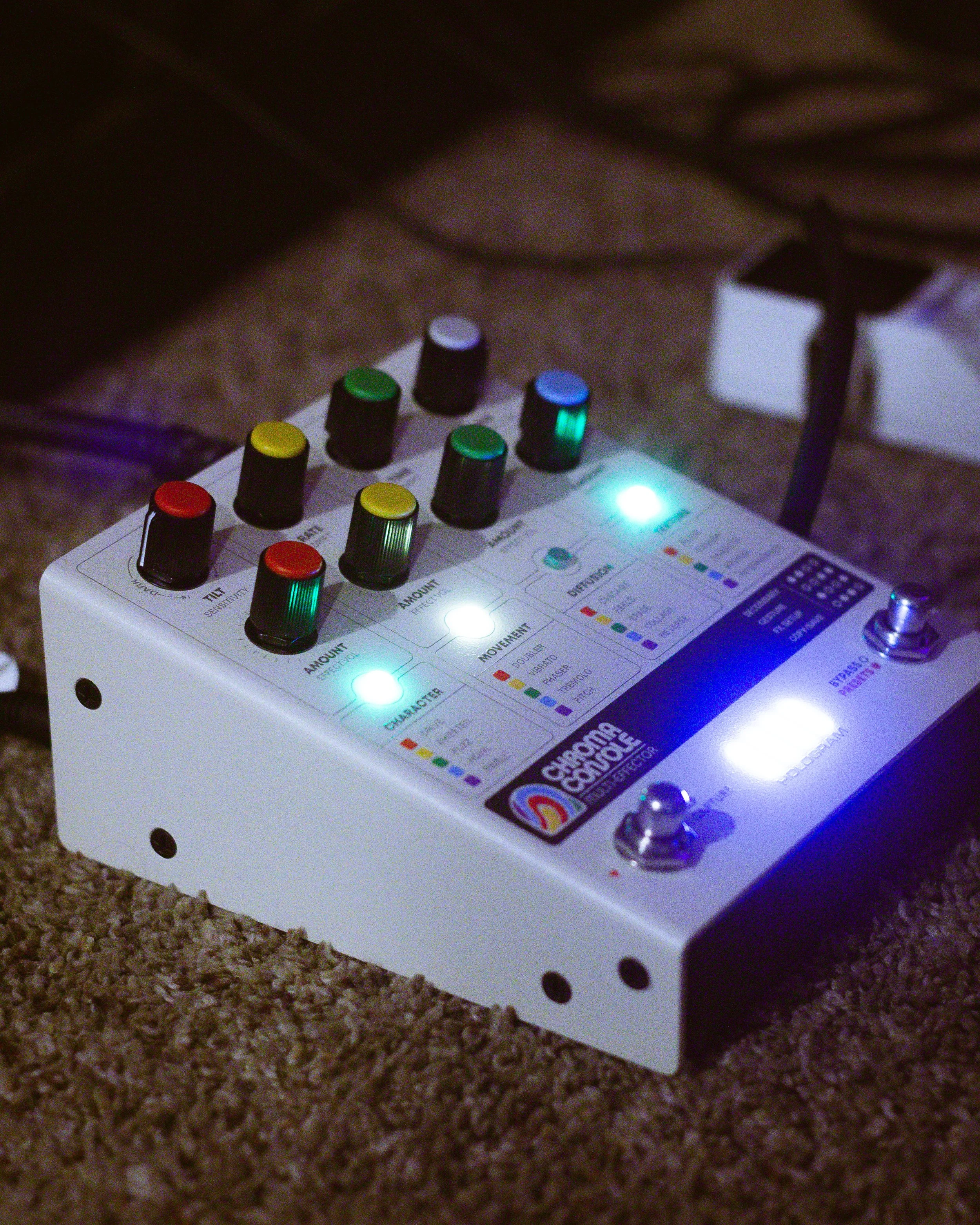 A white music effects pedal with colorful knobs, illuminated buttons, and various labels on a brown carpeted floor, connected to an electrical outlet.