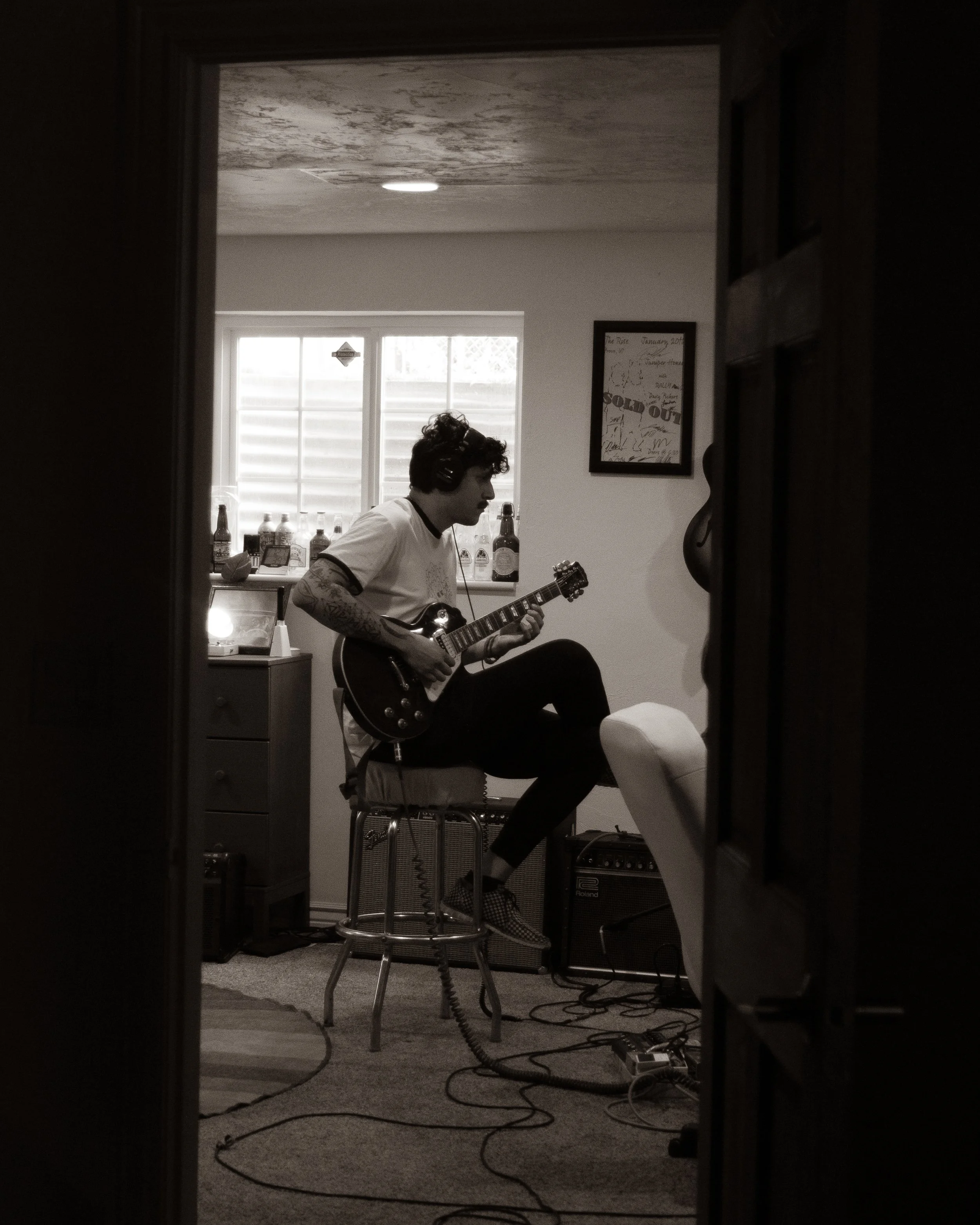 A young man with curly hair and tattoos playing an electric guitar in a cozy, dimly-lit room, seen through a doorway.