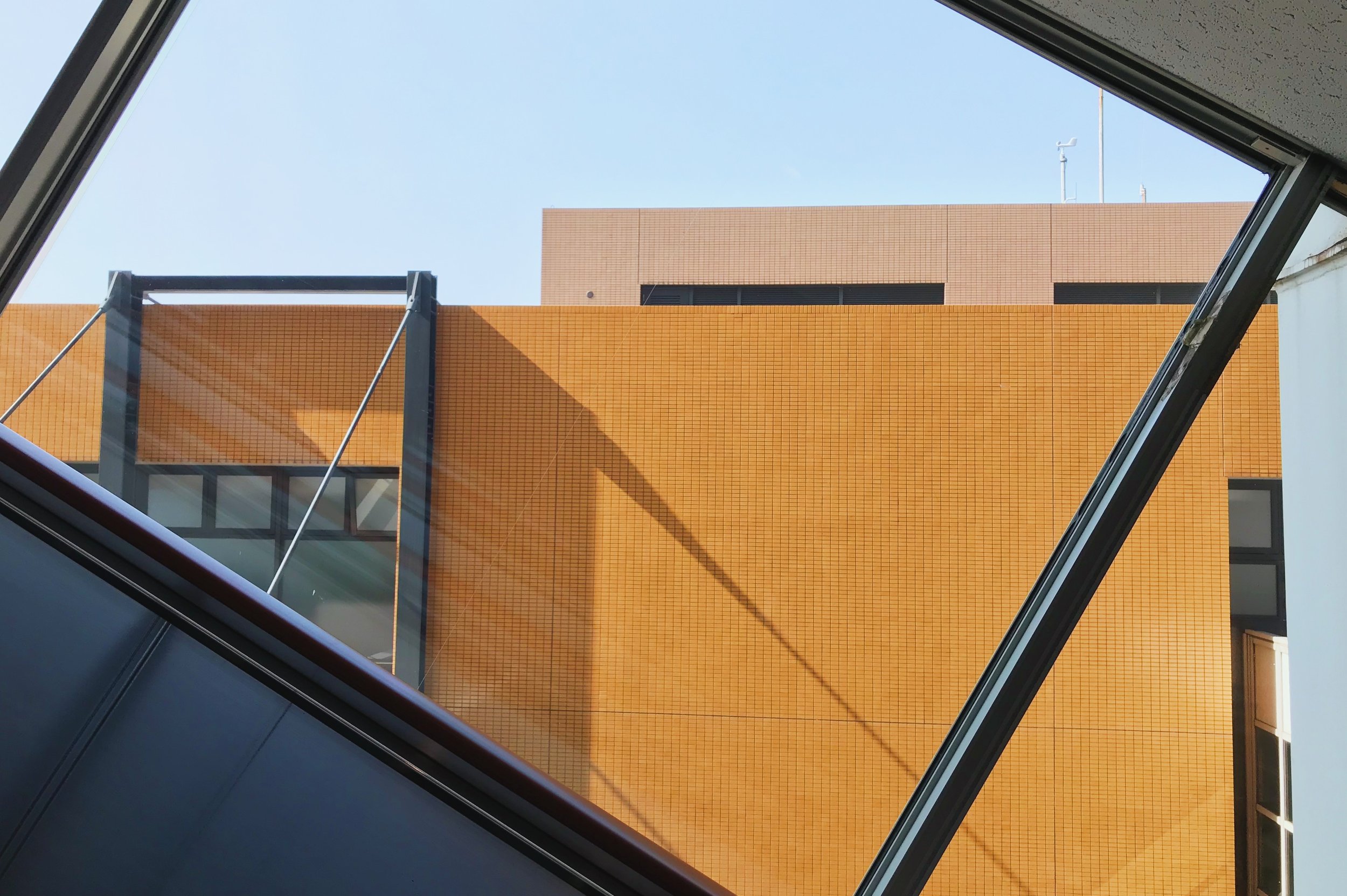 View of a modern building seen through an angled glass window from inside, with orange and beige exterior walls and clear blue sky.