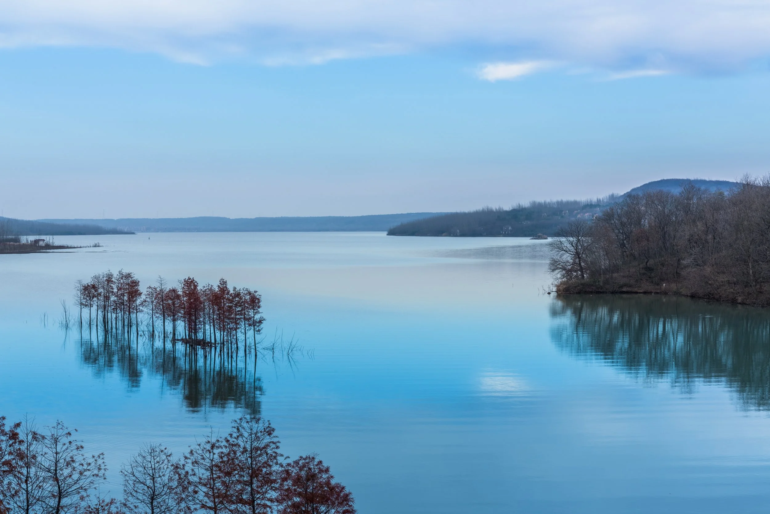 A serene body of water with a small cluster of trees in the foreground, with reflections on the water, and hills and trees in the background under a partly cloudy sky.