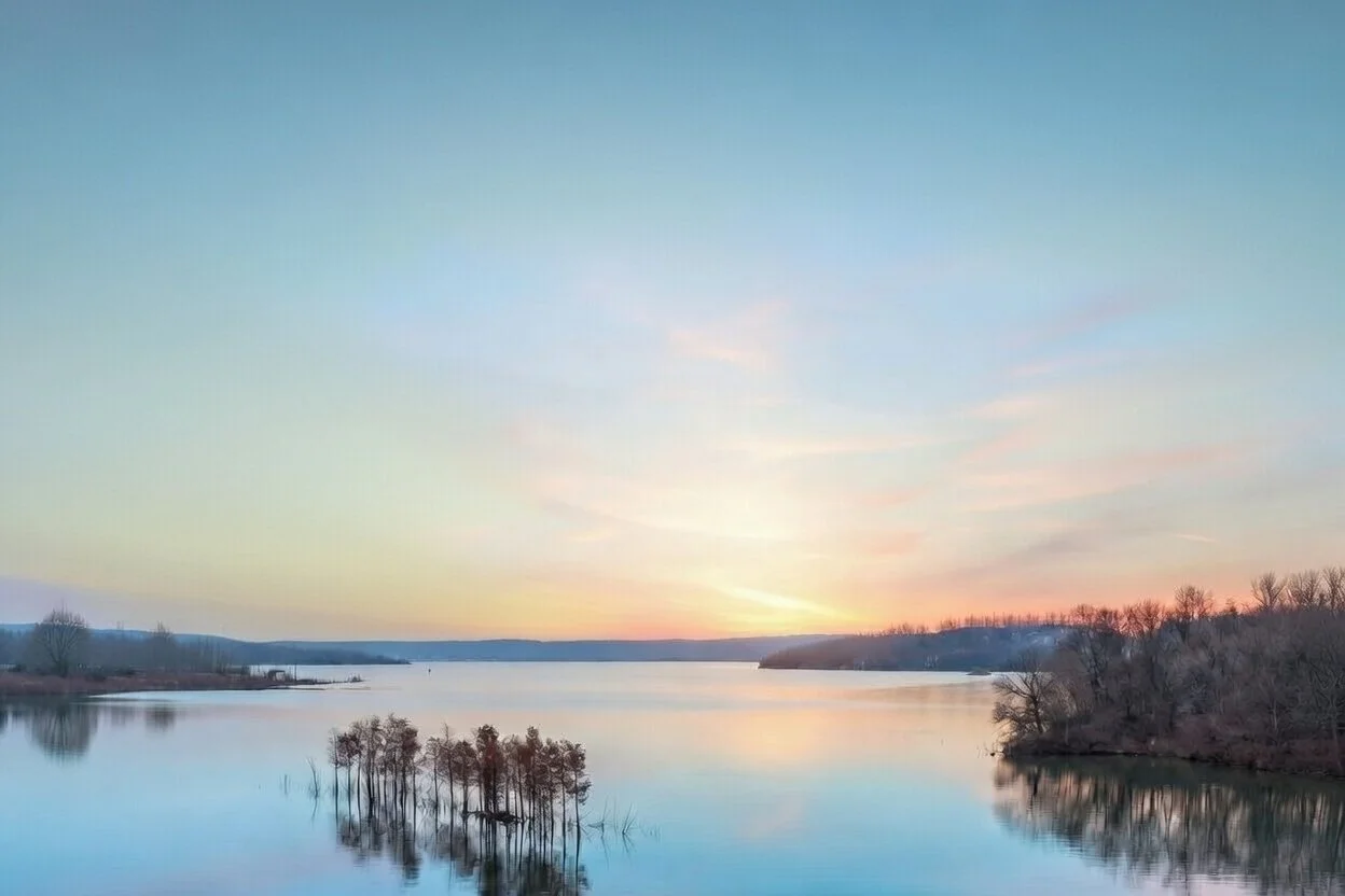 Serene lake at sunset with calm water reflecting distant trees and a pastel sky.