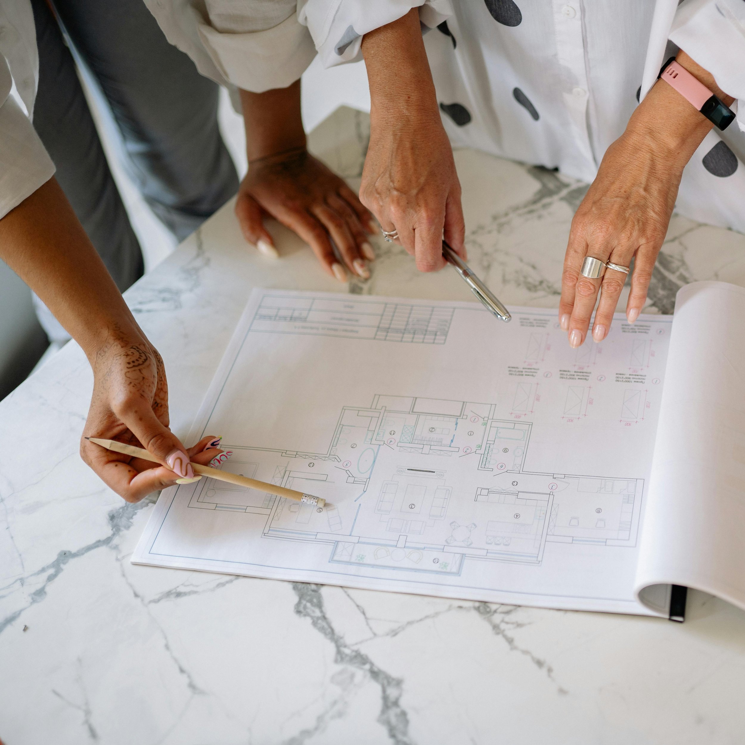 People gathered around a table looking at architectural plans and drawings, with one person holding a pencil and wearing a pink wristband.