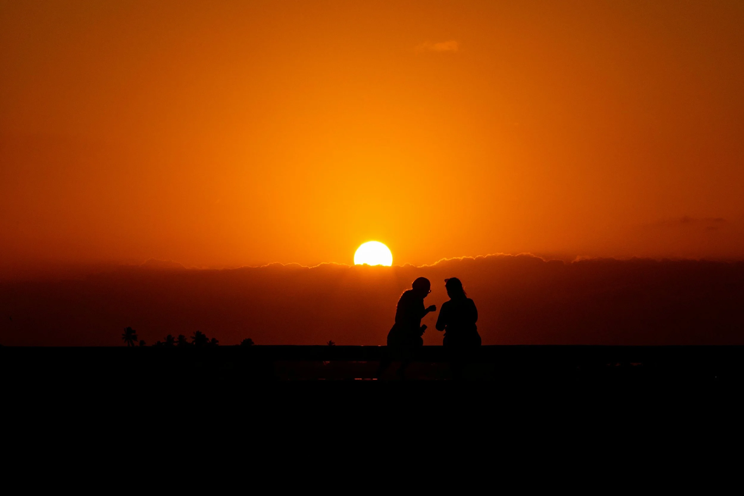 Silhouettes of two people talking during a sunset with the sun near the horizon and a dark sky, with trees in the distance.
