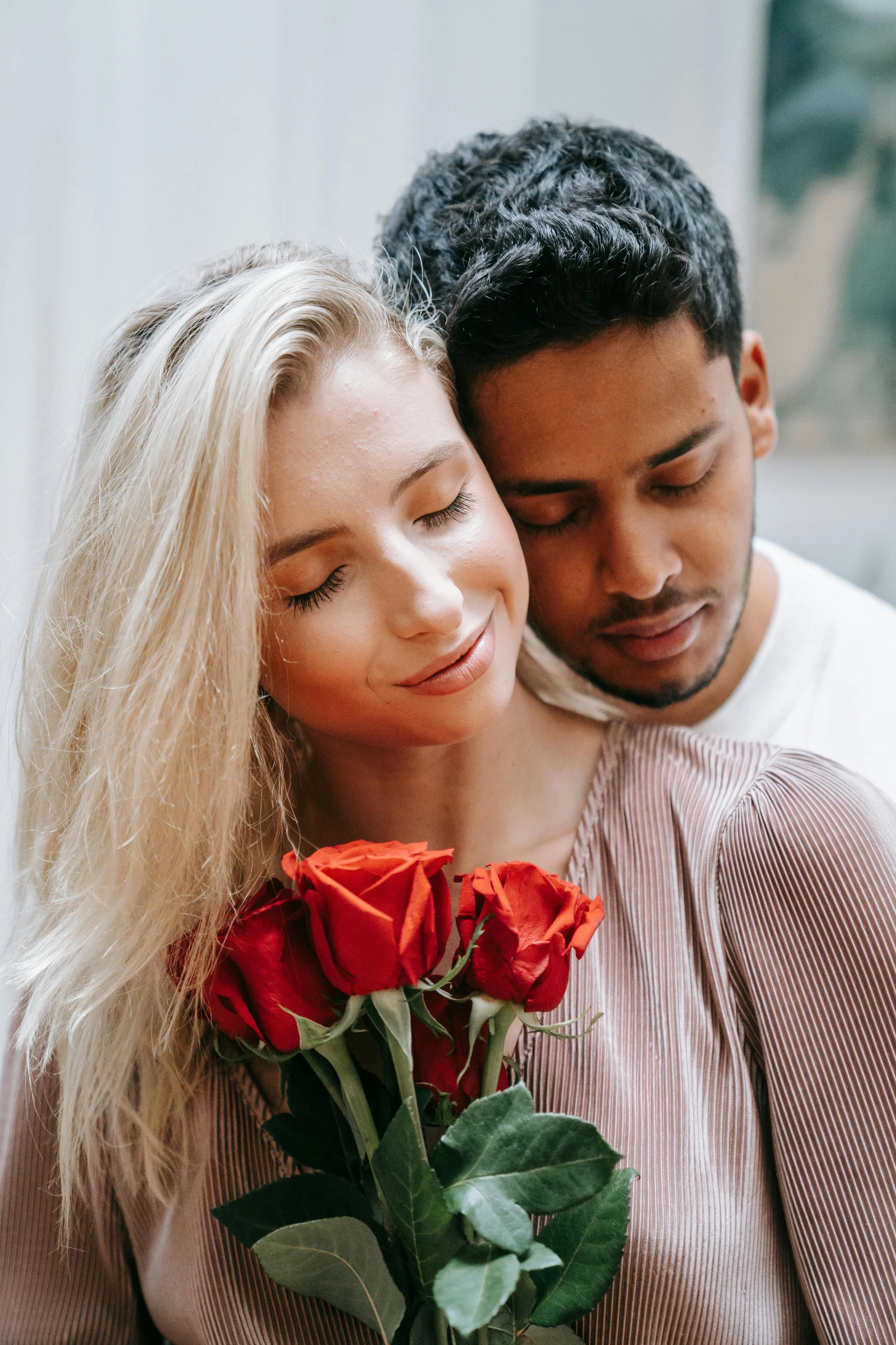 A couple with their eyes closed, smiling softly, holding a bouquet of red roses together.