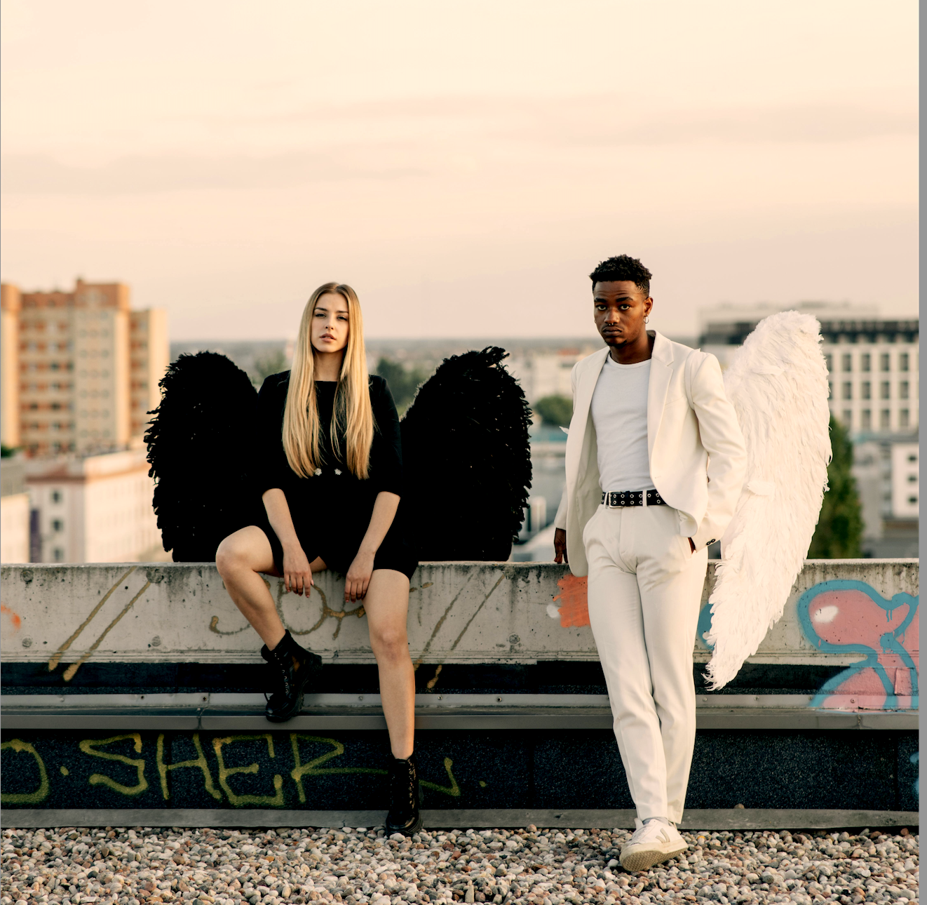 Two young people dressed as angels with wings stand on a rooftop with graffiti, city buildings in the background during sunset.