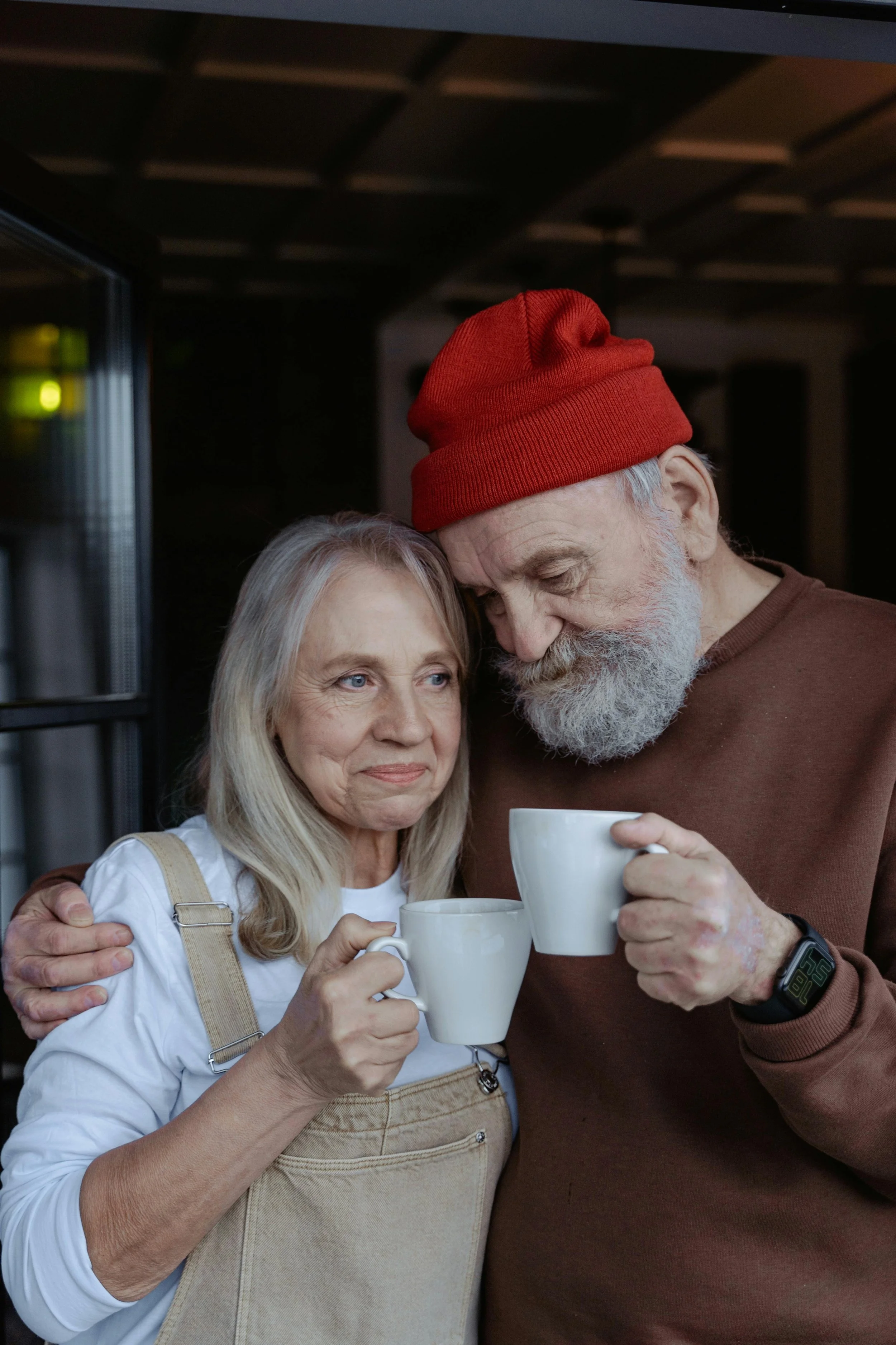 An elderly couple sharing a hug and clinking white coffee mugs in a cozy indoor setting. The woman has long gray hair, and the man has a gray beard and is wearing a red beanie and brown sweater.