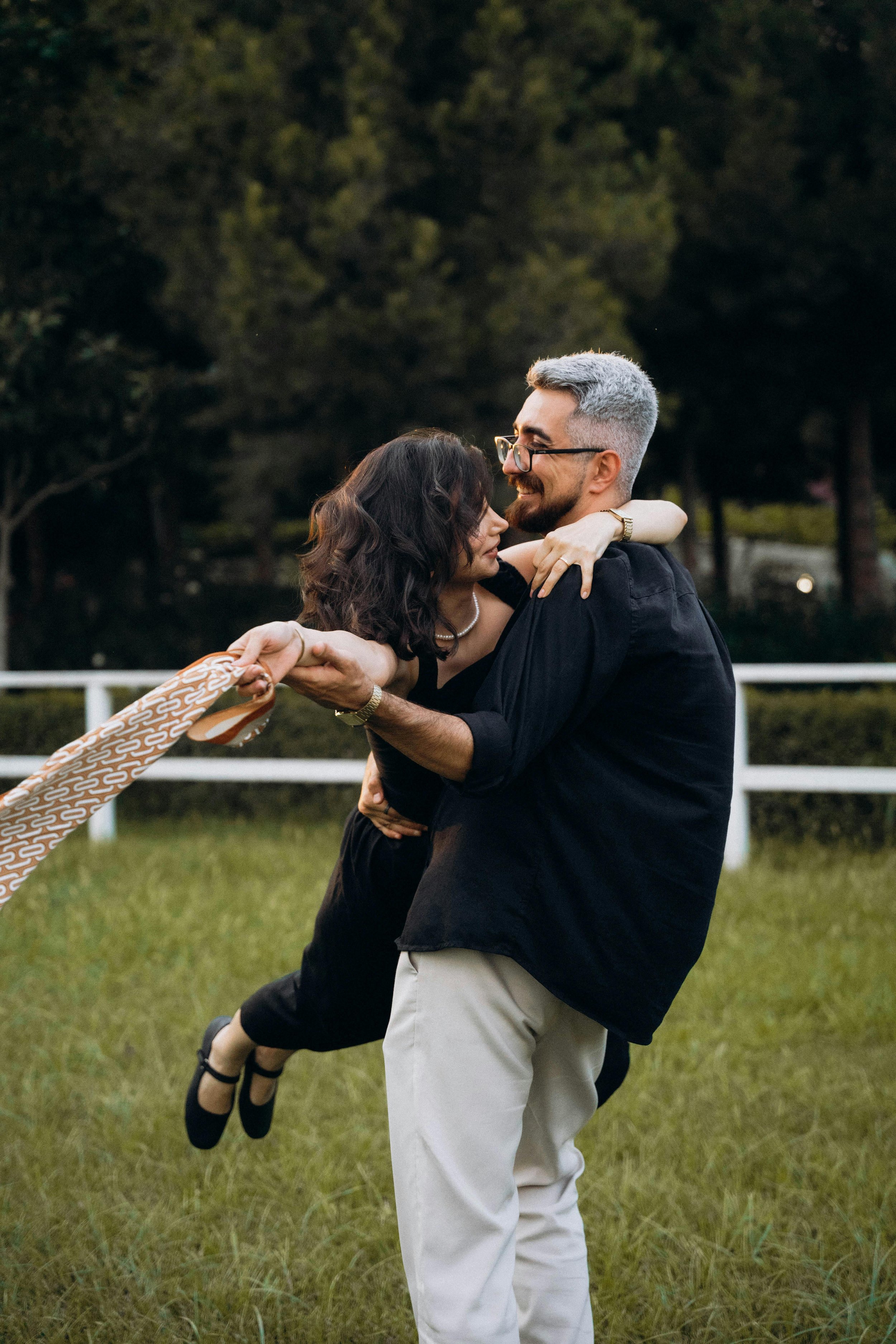A man holding a woman in a dance pose outdoors on a grassy field, with trees in the background, during what appears to be late evening or sunset.
