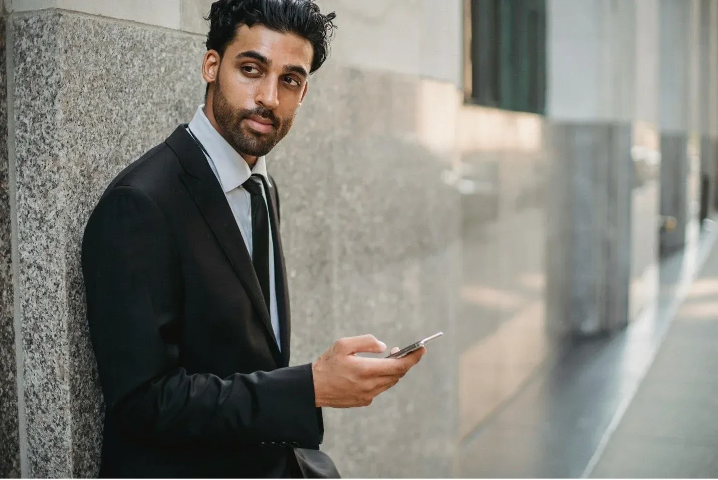 A man in a black suit and tie standing outside in front of a gray stone wall, looking to his left and holding a smartphone in his right hand.