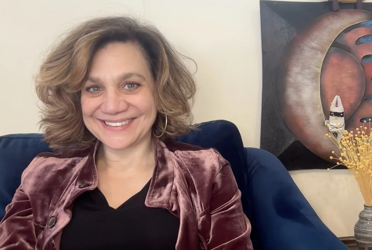 A woman with shoulder-length wavy hair smiling and sitting on a navy blue couch. Behind her, there is abstract artwork with large circular shapes and a wall with a light-colored background. To her right, a vase with yellow dried flowers is visible.