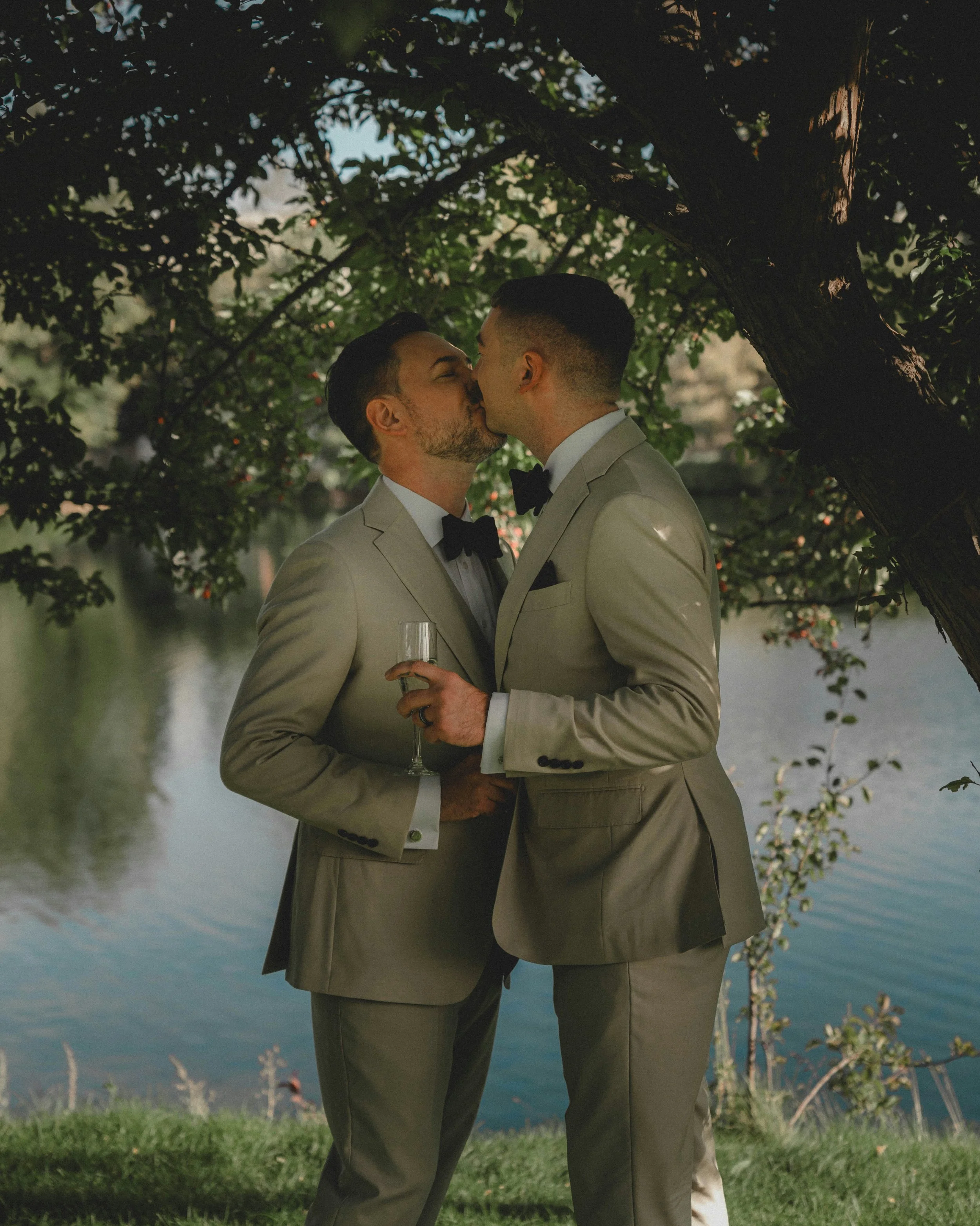 Two men in beige suits and black bow ties are kissing outdoors, one holding a champagne glass, beneath a tree by a body of water.