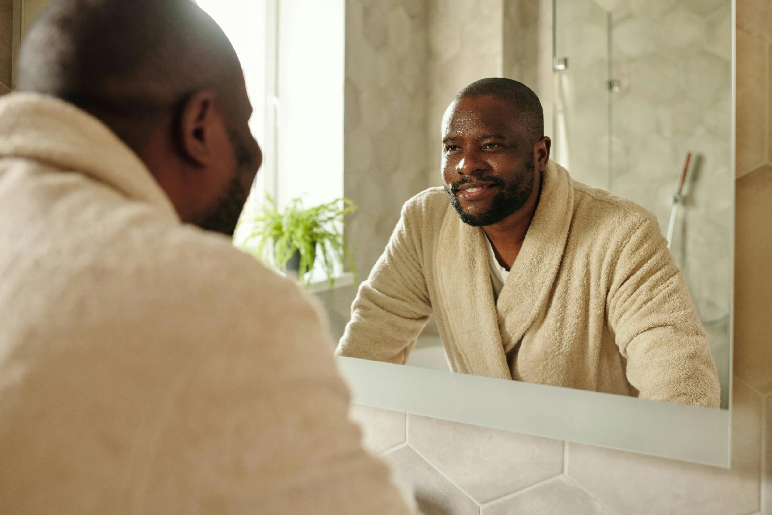 A man looking at himself in a bathroom mirror, smiling, wearing a beige bathrobe with natural light coming in from a window.