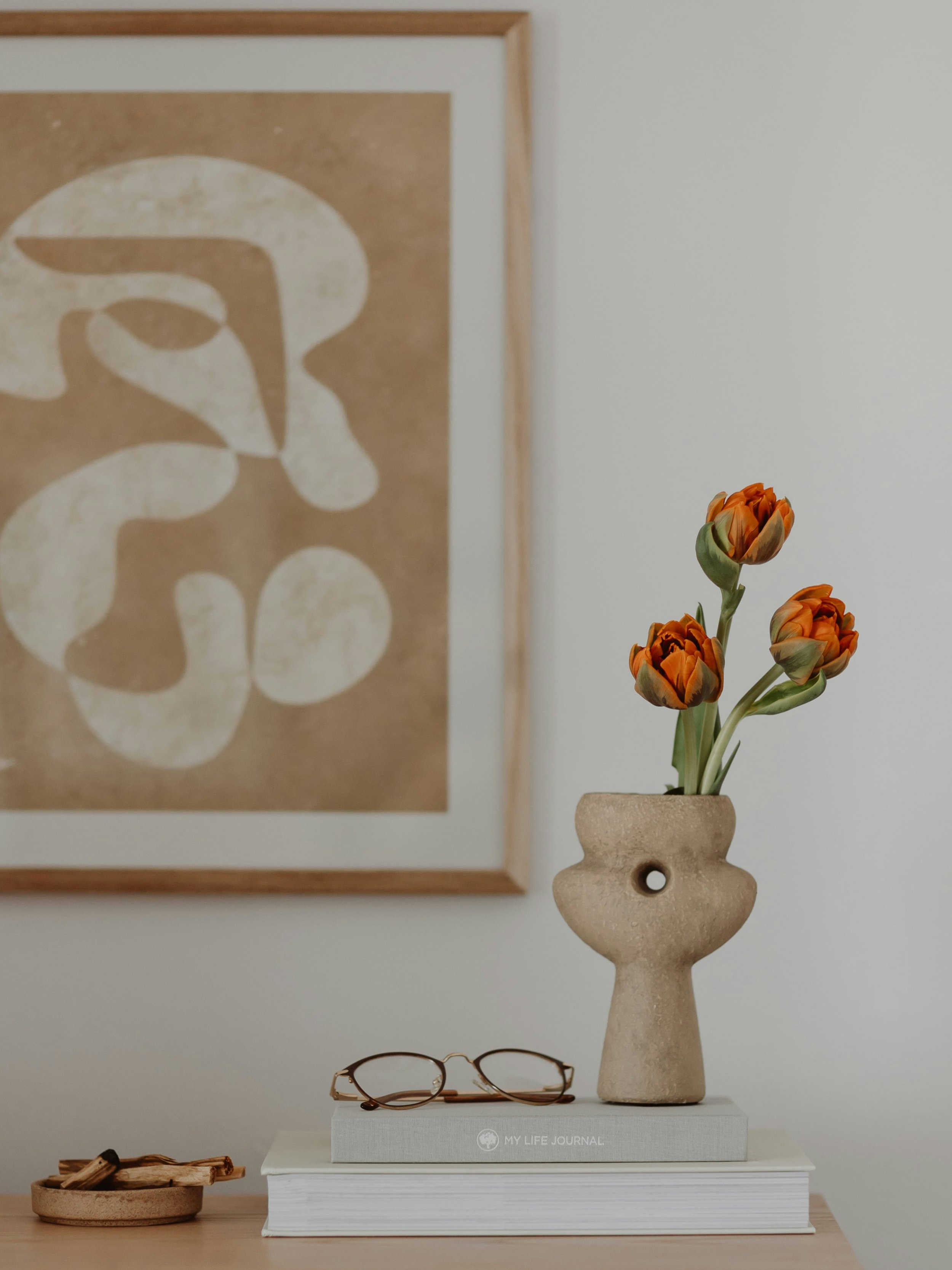 A beige vase with three orange flowers on a white book titled "My Life Journal" on a white surface, with glasses on the table and abstract artwork framed on the wall in the background.