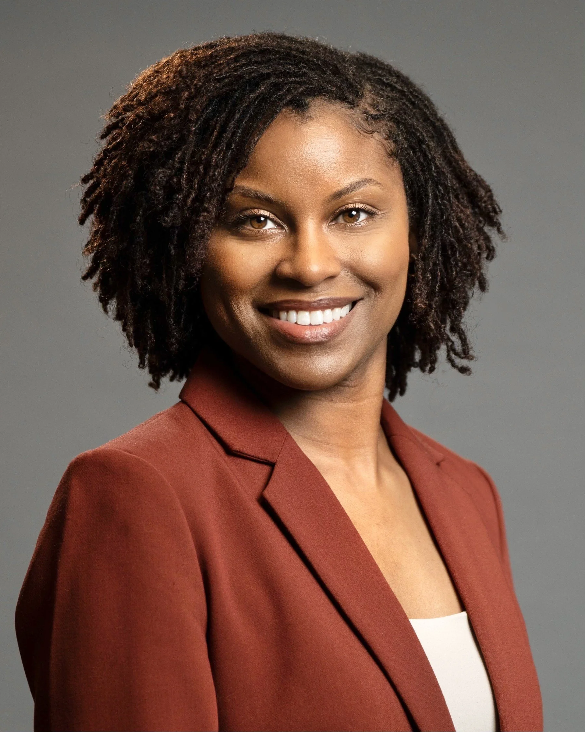 Headshot of a woman with curly hair wearing a rust-colored blazer and white top smiling at the camera against a gray background.