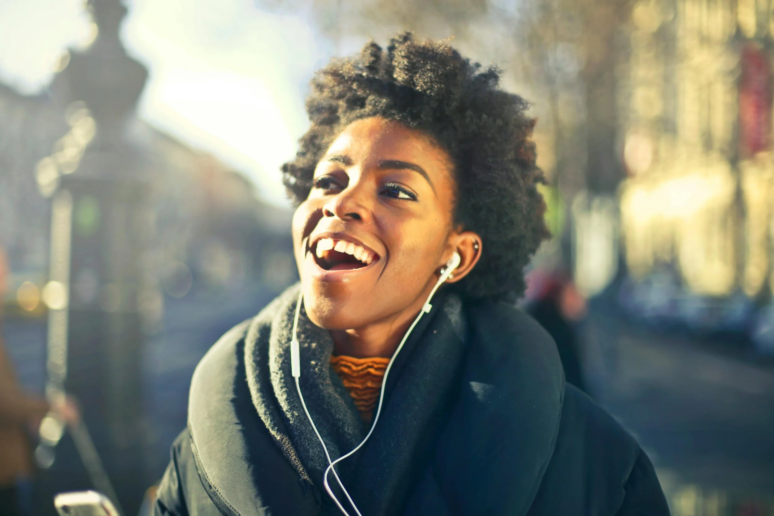 A young woman with curly hair, smiling and listening to music with earphones outdoors in sunny weather.