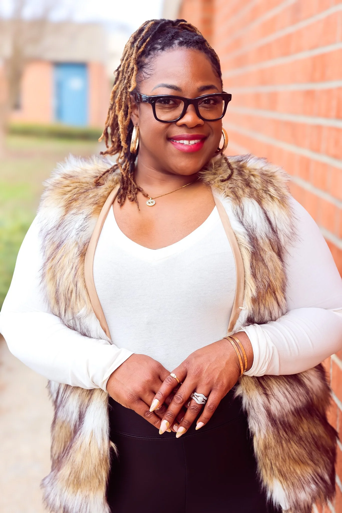 A woman with dark skin and dreadlocks wearing glasses, gold hoop earrings, and a gold necklace standing outdoors against a brick wall. She is dressed in a white top, a fur vest, and black pants, smiling and posing for the camera.