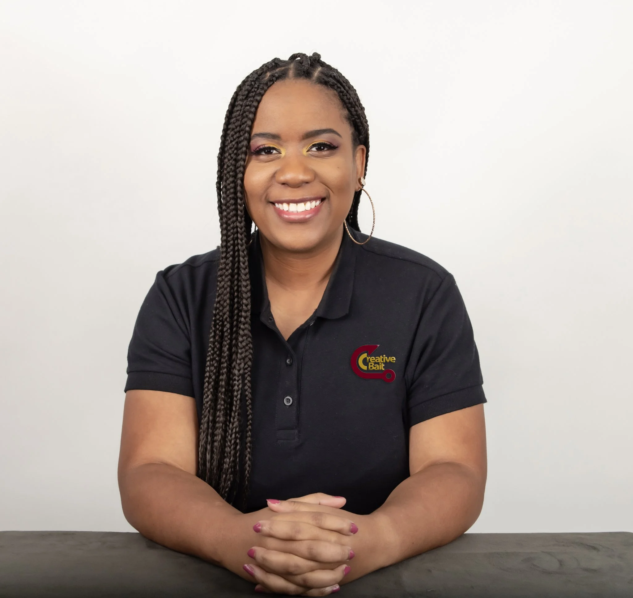 A woman with long braids, wearing hoop earrings and a black polo shirt with a logo that reads 'Creative Bait,' smiling while sitting at a table against a plain white background.
