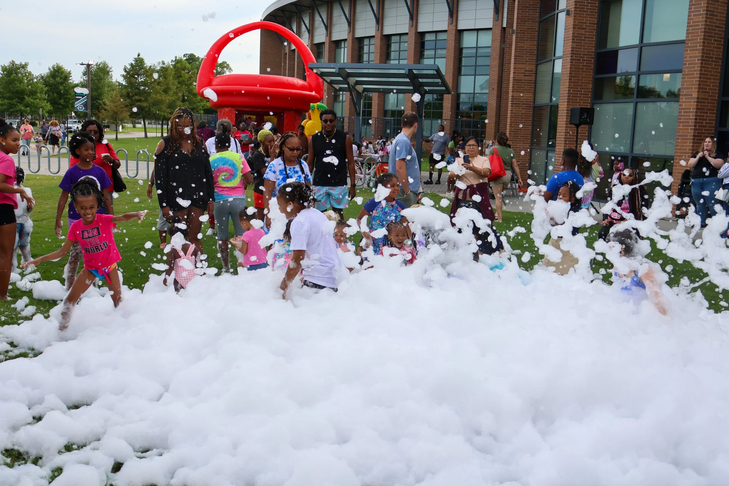 Children and adults playing and having fun in foam outdoors in front of a brick building with large glass windows.